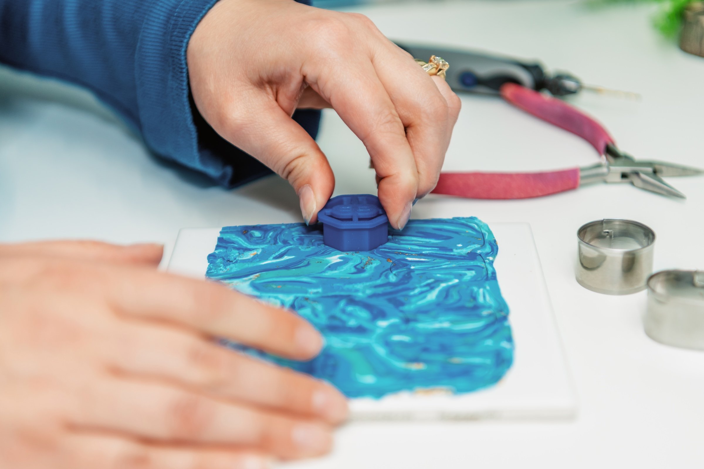 A person pressing a blue seal onto a piece of blue, marbled clay or polymer with their hand. There are jewelry-making tools and metal cookie cutters on the table, indicating a craft or jewelry-making activity.