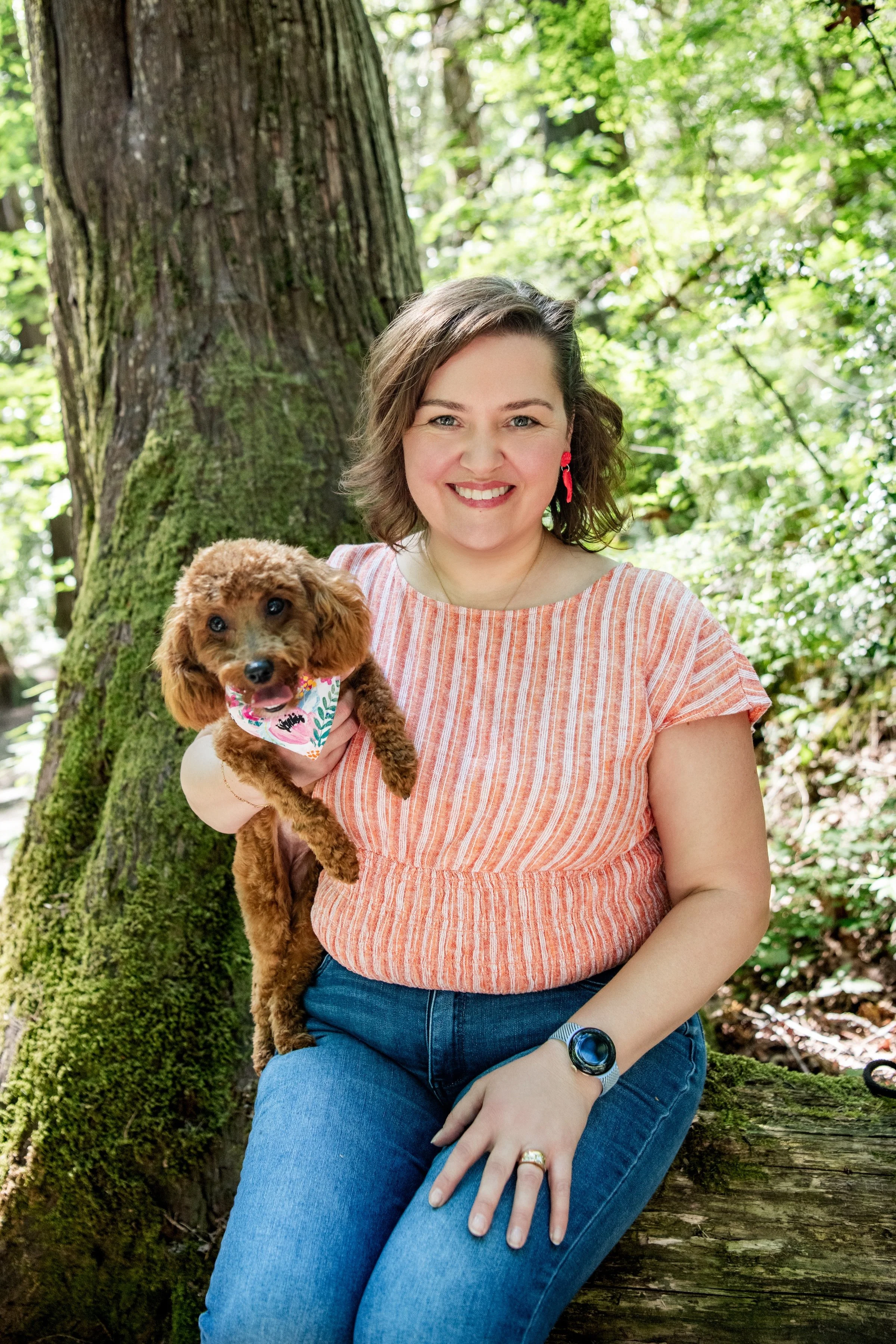 A woman with short brown hair and red earrings holding a small brown dog wearing a colorful bandana, sitting by a large tree in a sunny forest.
