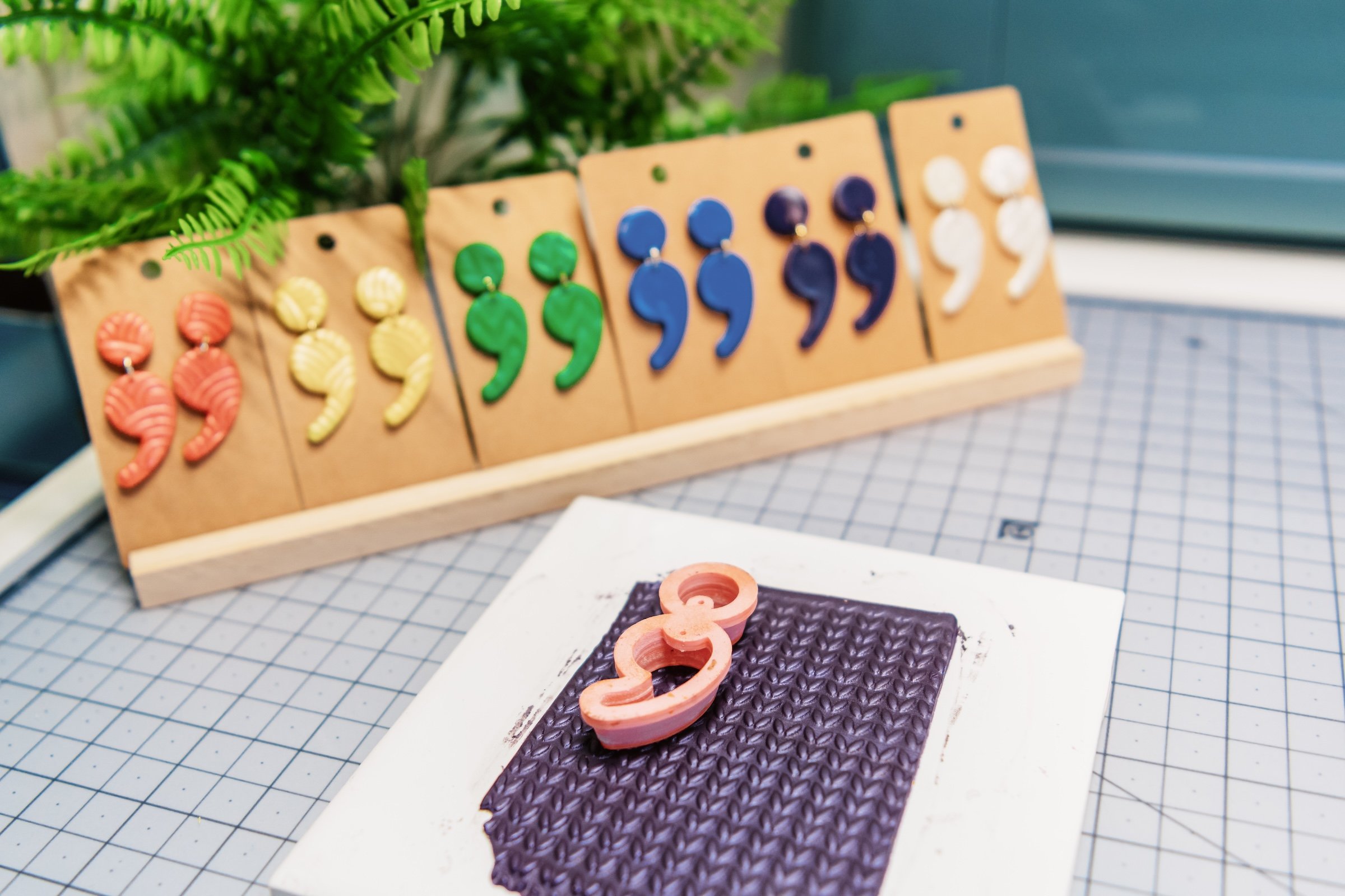Colorful earrings displayed on a wooden stand, with fern plants in the background and a textured chocolate-colored surface with the number three on it in the foreground.