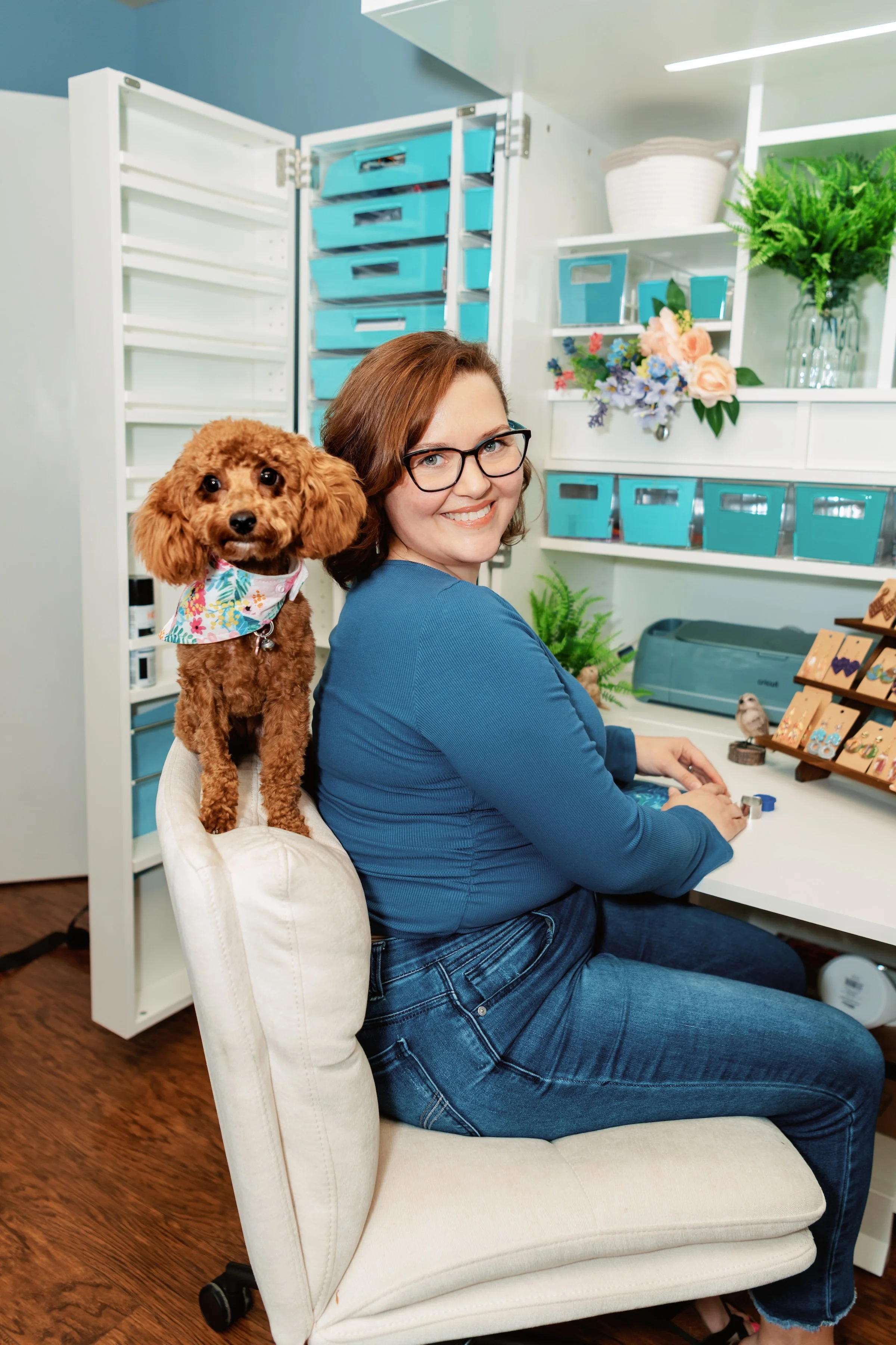 A woman with glasses and a blue shirt smiling while sitting at a desk with a small poodle dog on her shoulder. The dog is wearing a floral bandana and looking at the camera. There is a white chair, open storage cabinets with blue bins, flowers, and office supplies in the background.