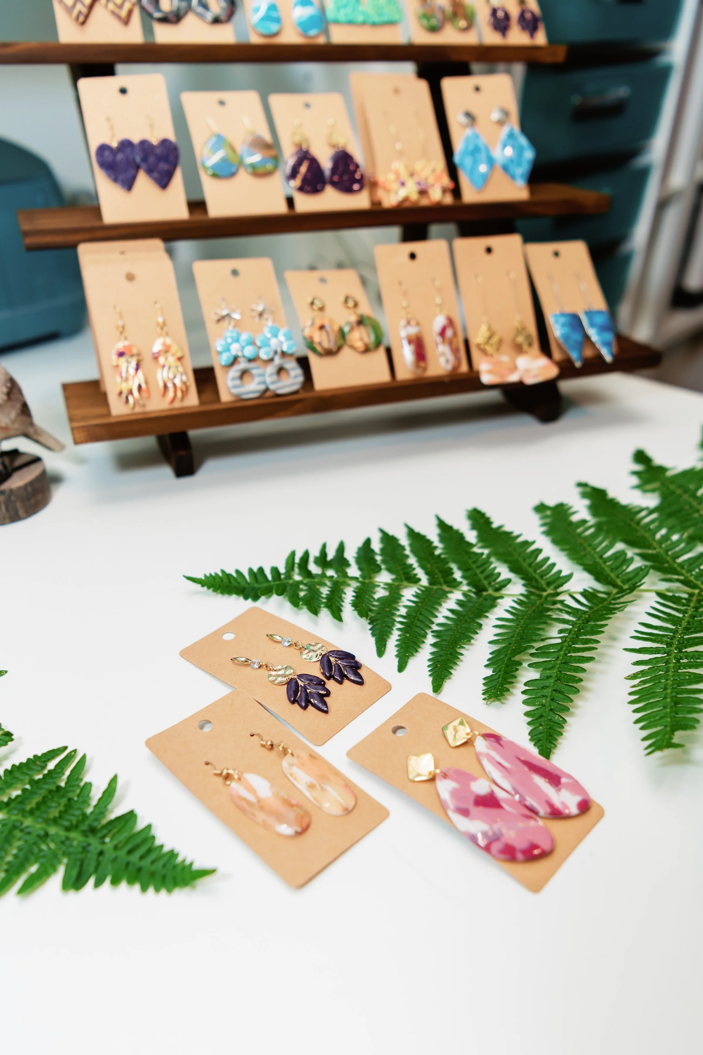 Display of colorful earrings on beige cards, some with floral motifs, arranged on a wooden stand with greenery in the foreground.