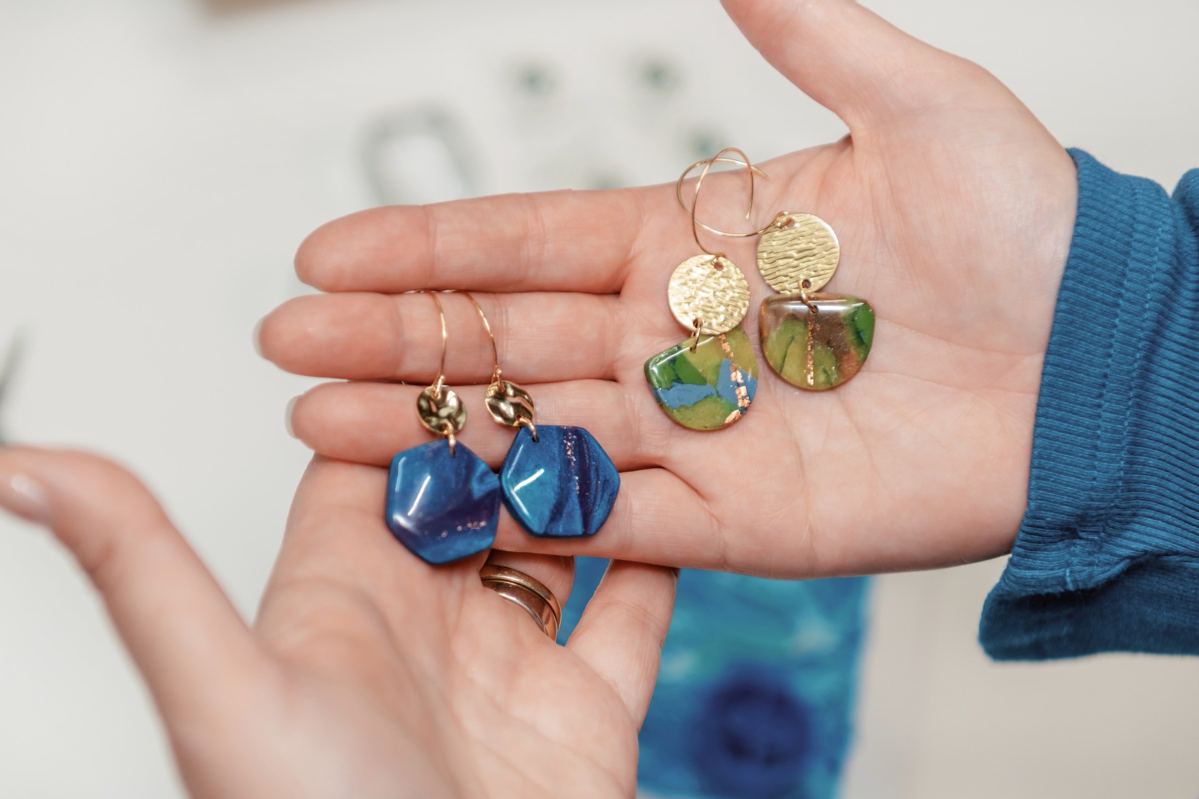 Close-up of a person's hand holding colorful earrings, with a white background and blurred jewelry displays in the background.