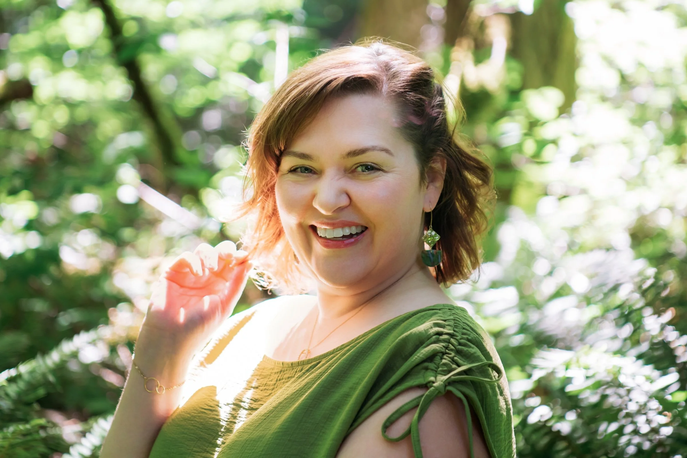 A woman with shoulder-length reddish hair smiling in a green dress, standing outdoors among trees with sunlight filtering through the leaves.