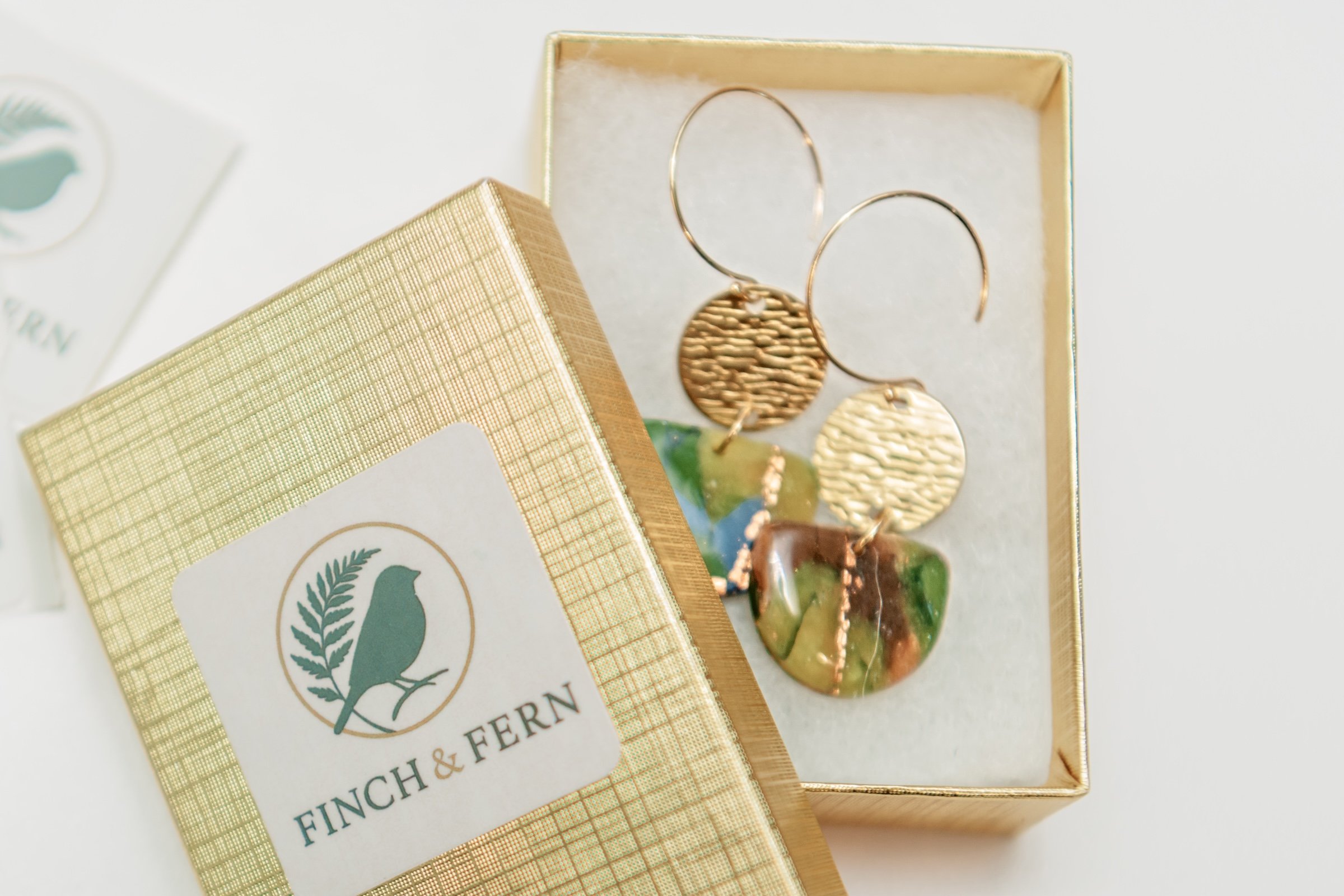 Gold box containing a pair of gold hoop earrings with circular charms and a marbled soap bar with green, brown, and cream colors.