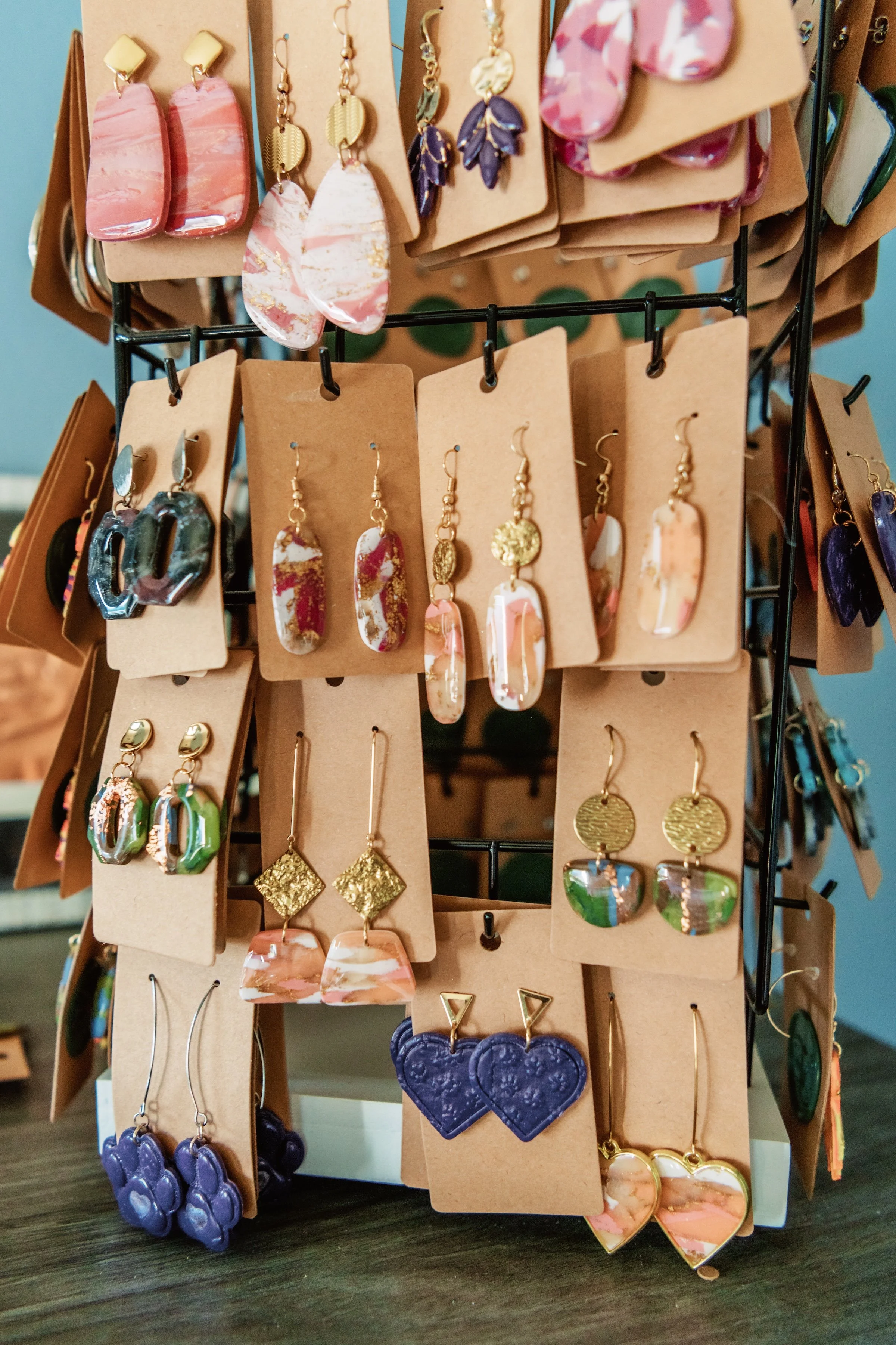 Display of various earrings on brown cardboard cards arranged on a black metal stand. The earrings include different styles such as pink and purple abstract shapes, gold circles, geometric gold and marble designs, and heart-shaped designs in purple and pink.