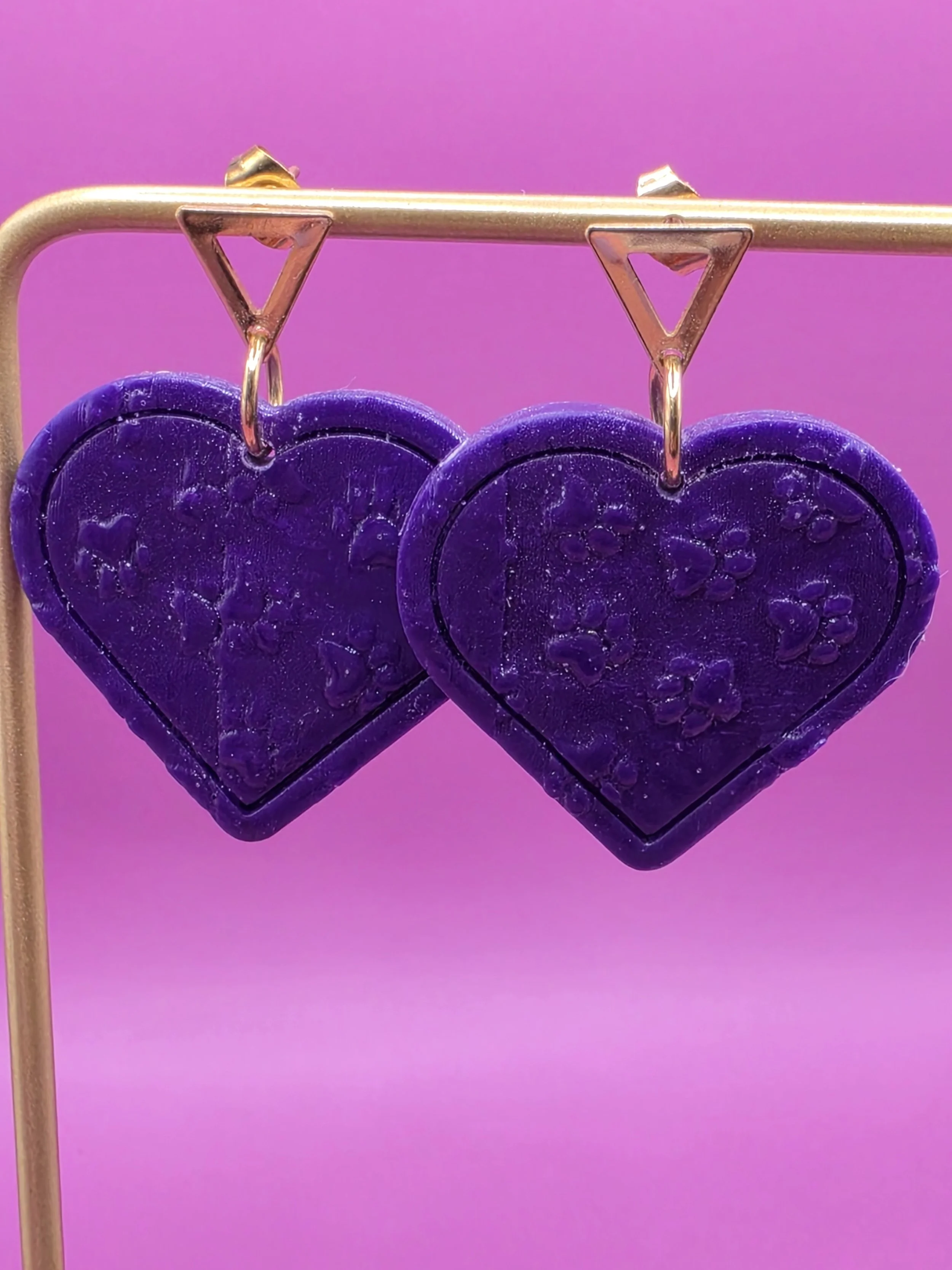 Purple heart-shaped earrings hanging from a gold wire against a pink background.