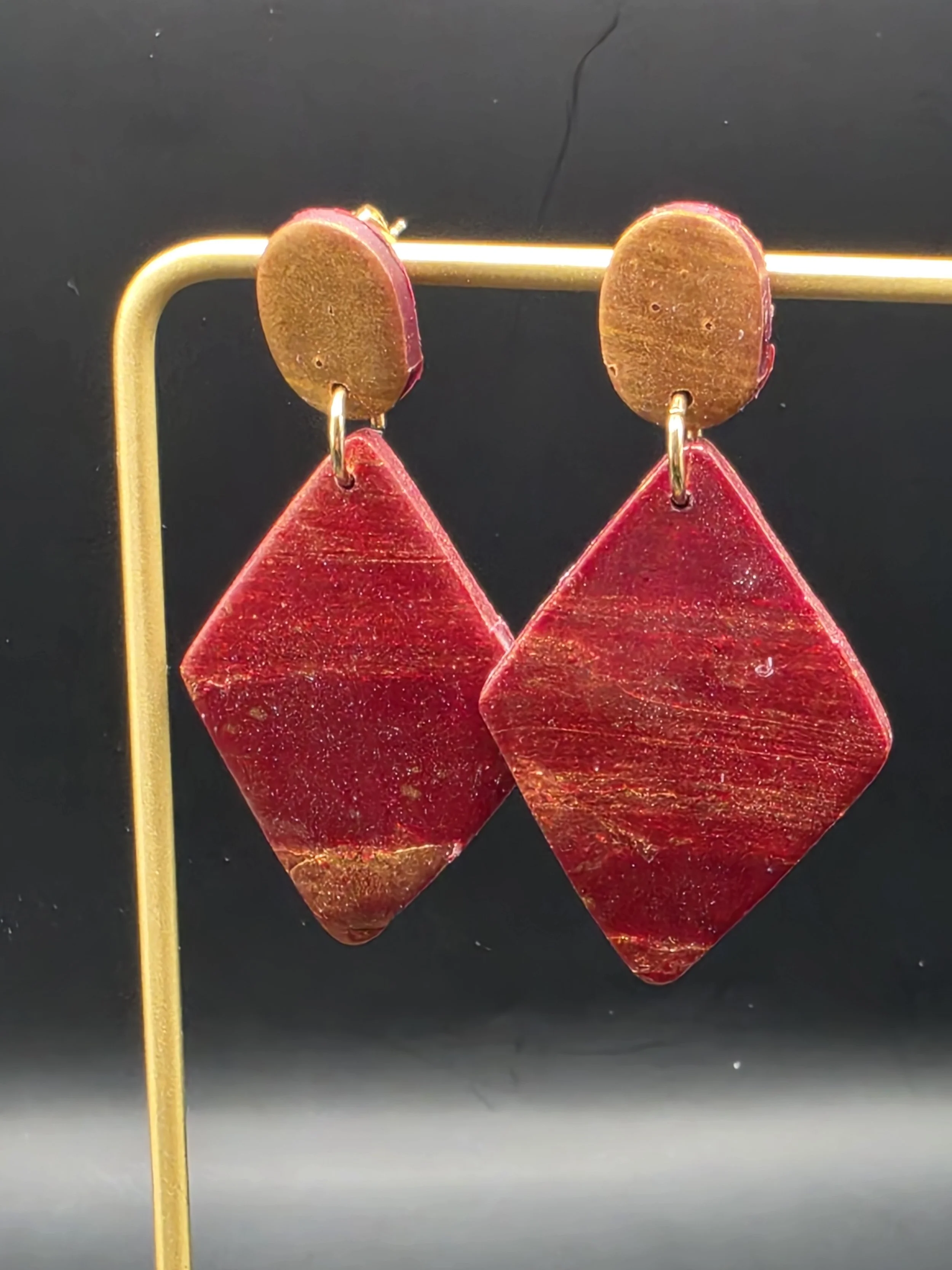 Close-up of red and gold earrings with geometric shapes hanging from a gold rod against a dark background.