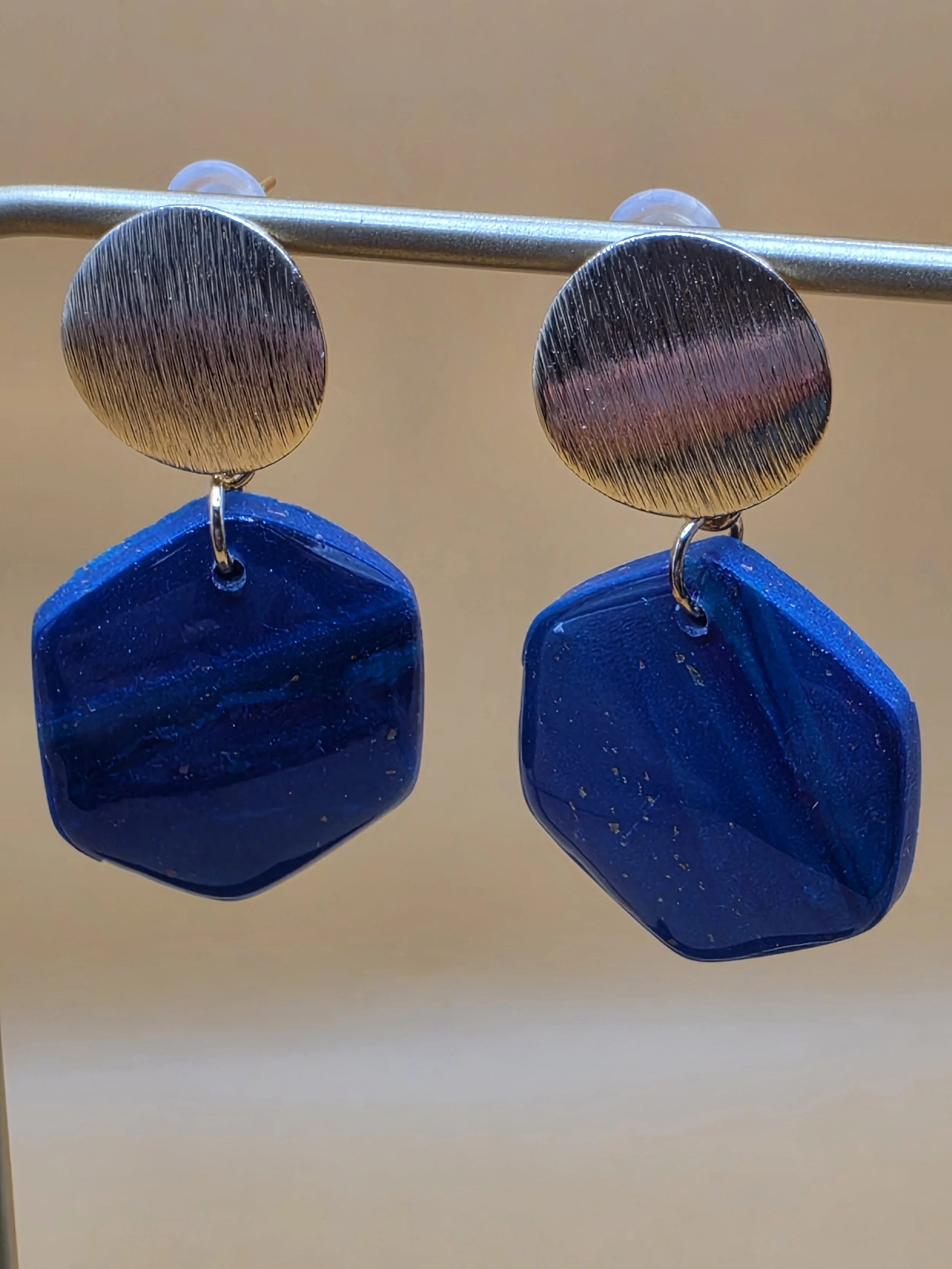 Close-up of a pair of dangling earrings with a round, textured metallic top and a large, faceted blue gemstone at the bottom, hanging from a metal rod against a plain beige background.