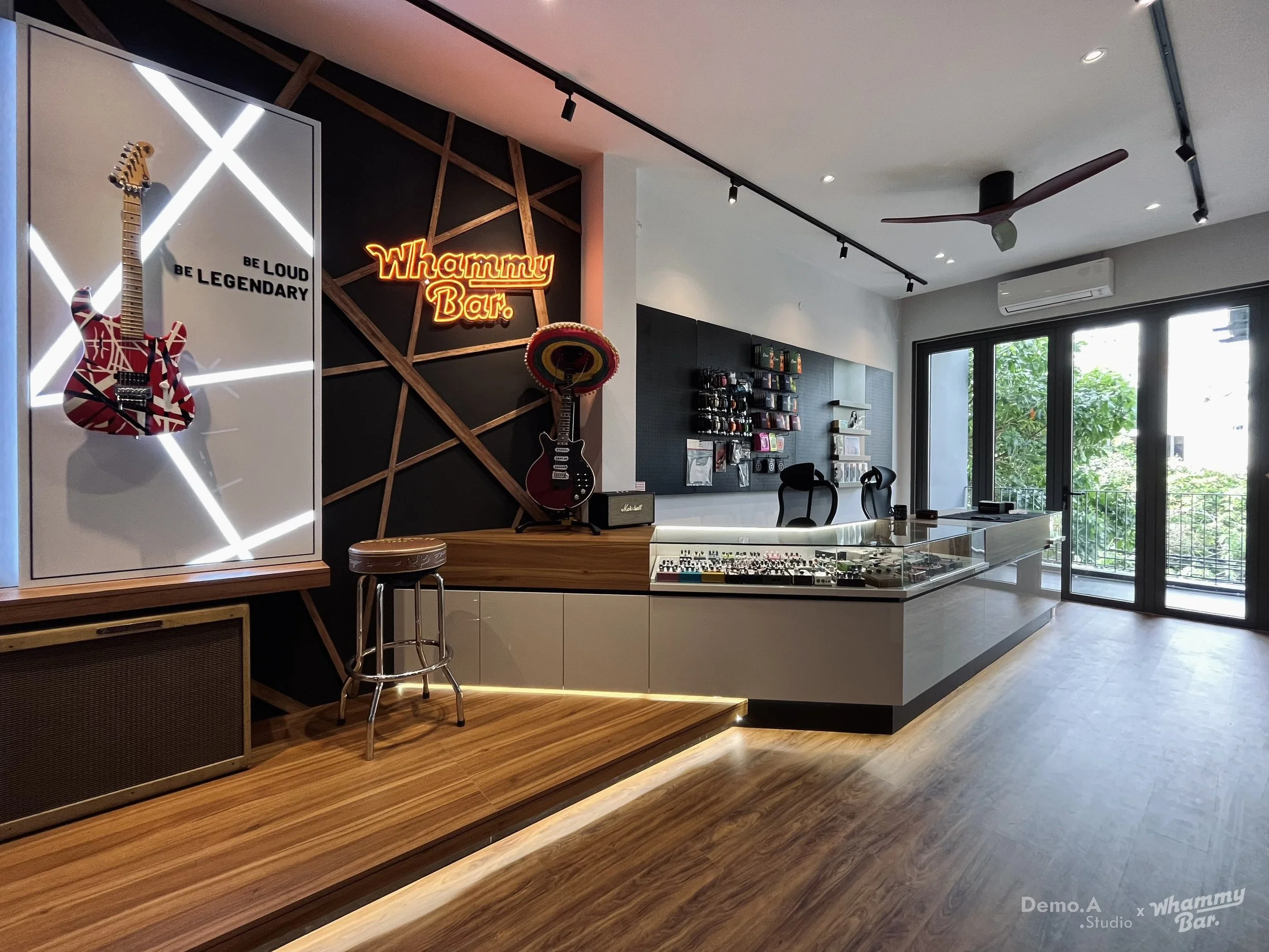 Interior of a modern music shop with guitars on display, a neon sign that reads 'Whammy Bar,' shelves with music accessories, and large windows letting in natural light.