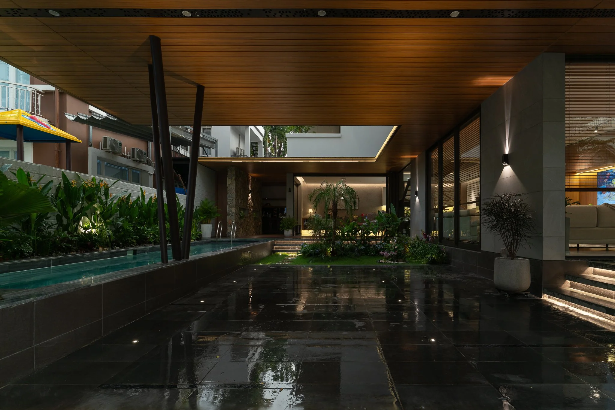 Modern outdoor lounge area with a wooden ceiling, glass walls, lush plants, and a reflecting pool.