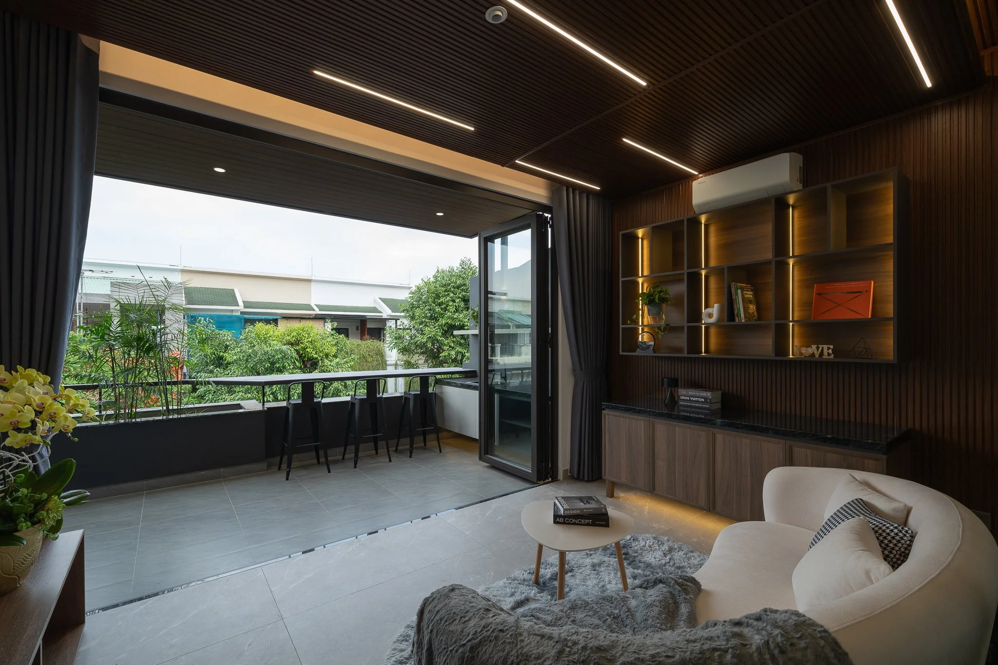 Living room with large open window, gray curtains, wooden bookshelves with decor, and a balcony with seating. Interior includes a beige sofa with black and white pillows, small round coffee table, plants, and ceiling lighting.