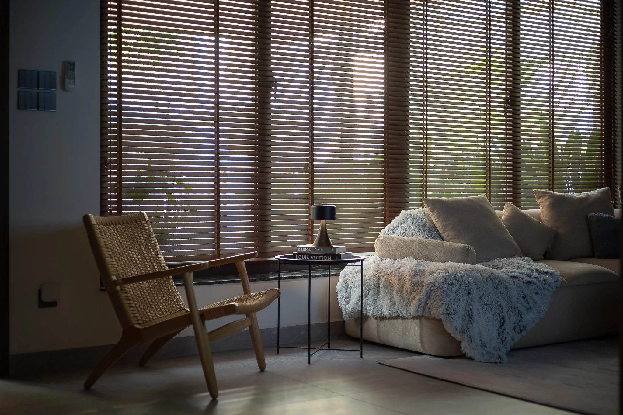 Living room with large windows covered by wooden blinds, a sofa with pillows and a fluffy throw blanket, a black table with books and a small black lamp, a woven chair, and a white wall with thermostat and light switch.