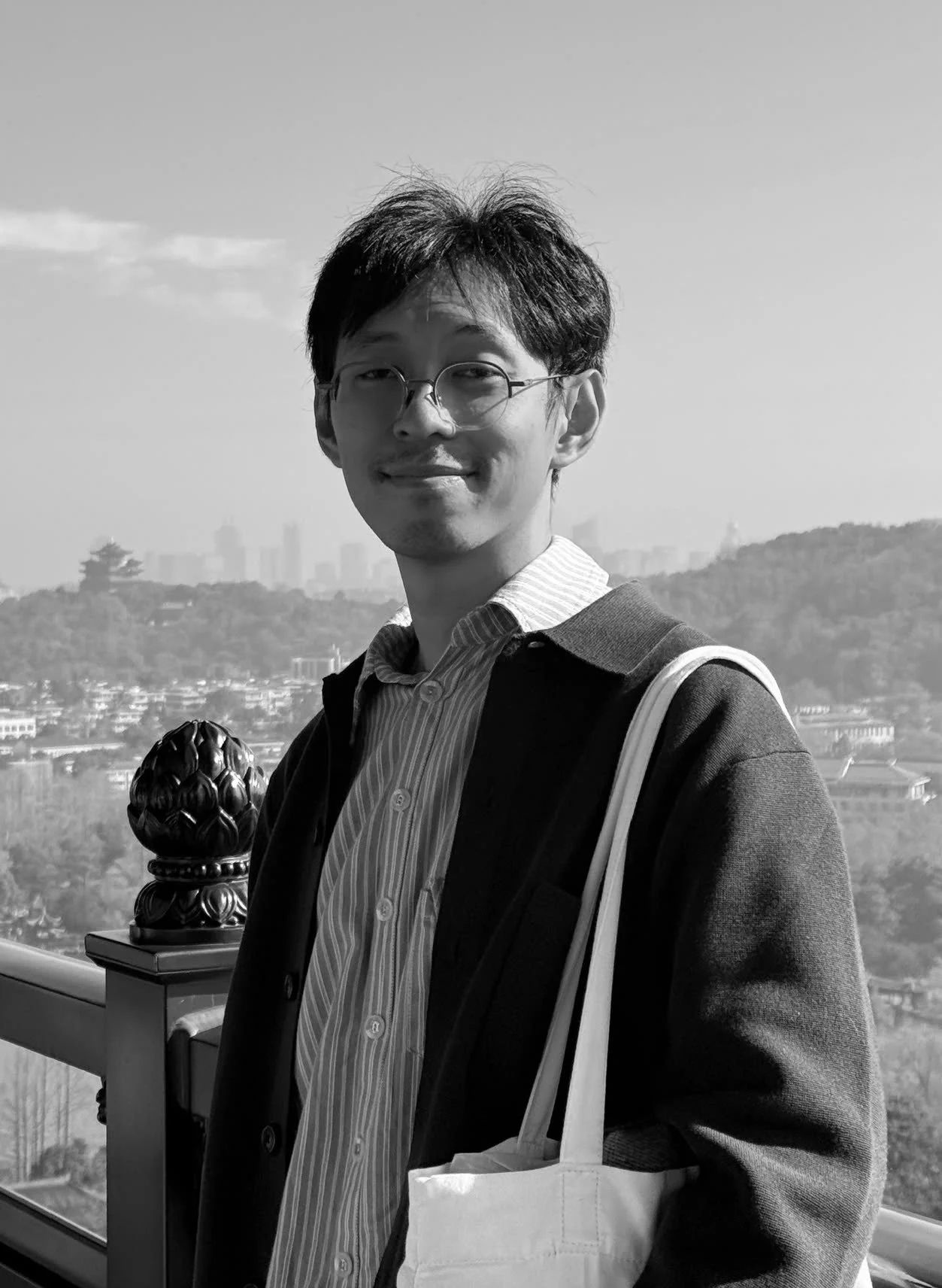 A black and white photo of a young man with glasses standing outdoors on a balcony or terrace, with a cityscape and hills in the background, wearing a striped shirt and a jacket, carrying a tote bag over his shoulder, smiling.