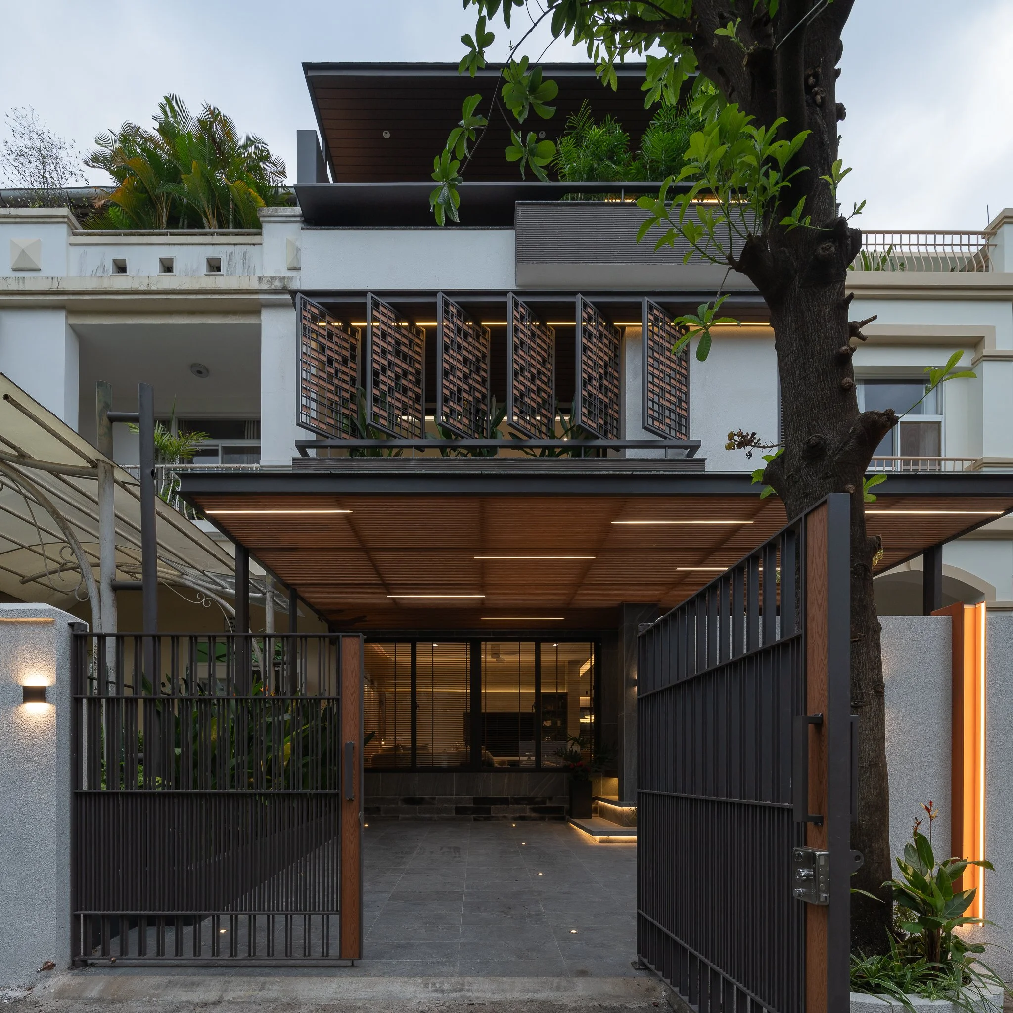 Modern residential entrance with metal gate, open wooden ceiling, and a staircase leading inside, framed by a large tree and greenery.