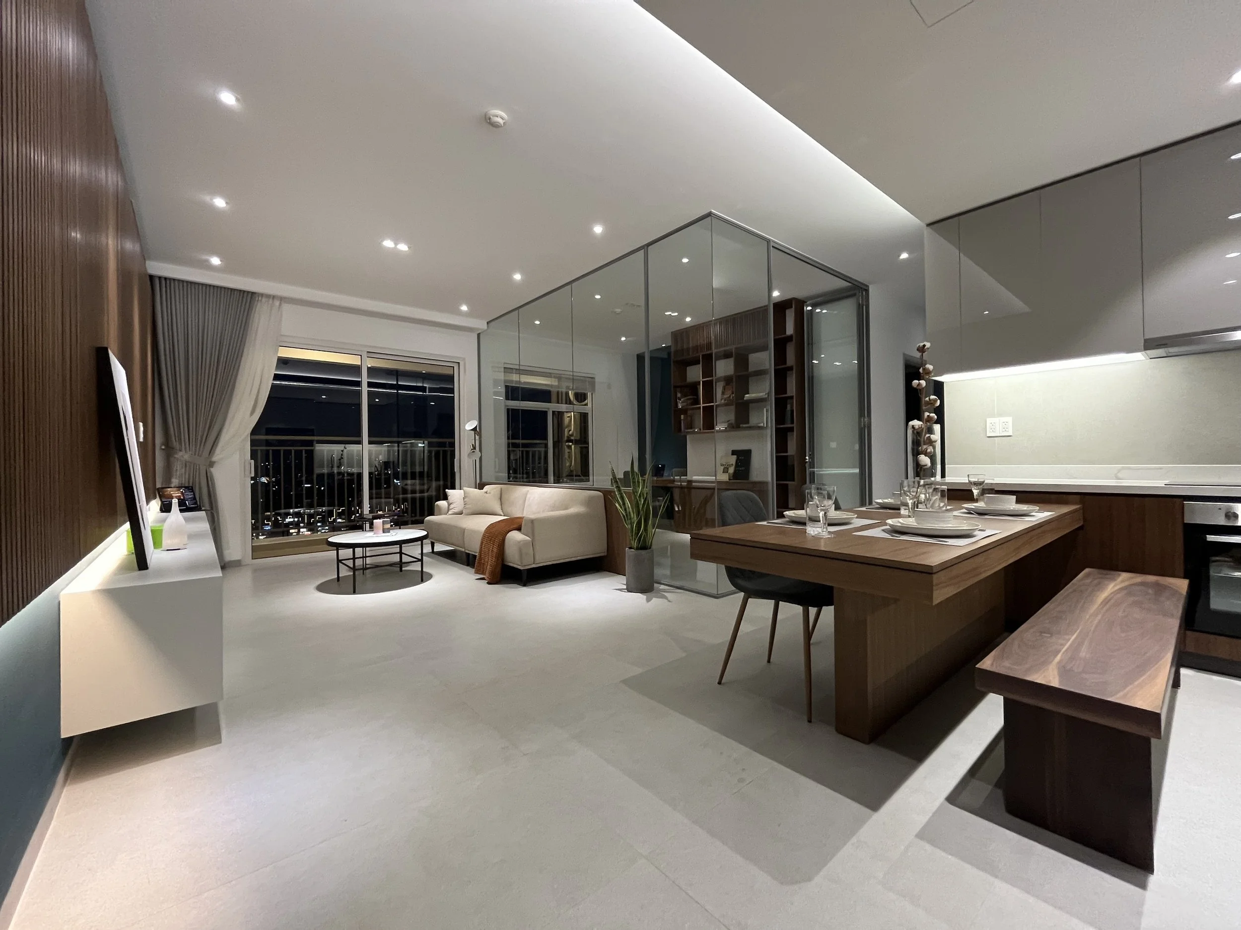 Modern living room and dining area with beige sofa, white curtains, hardwood bookshelf, glass partition, kitchen counter with table setting, and lighting fixtures.