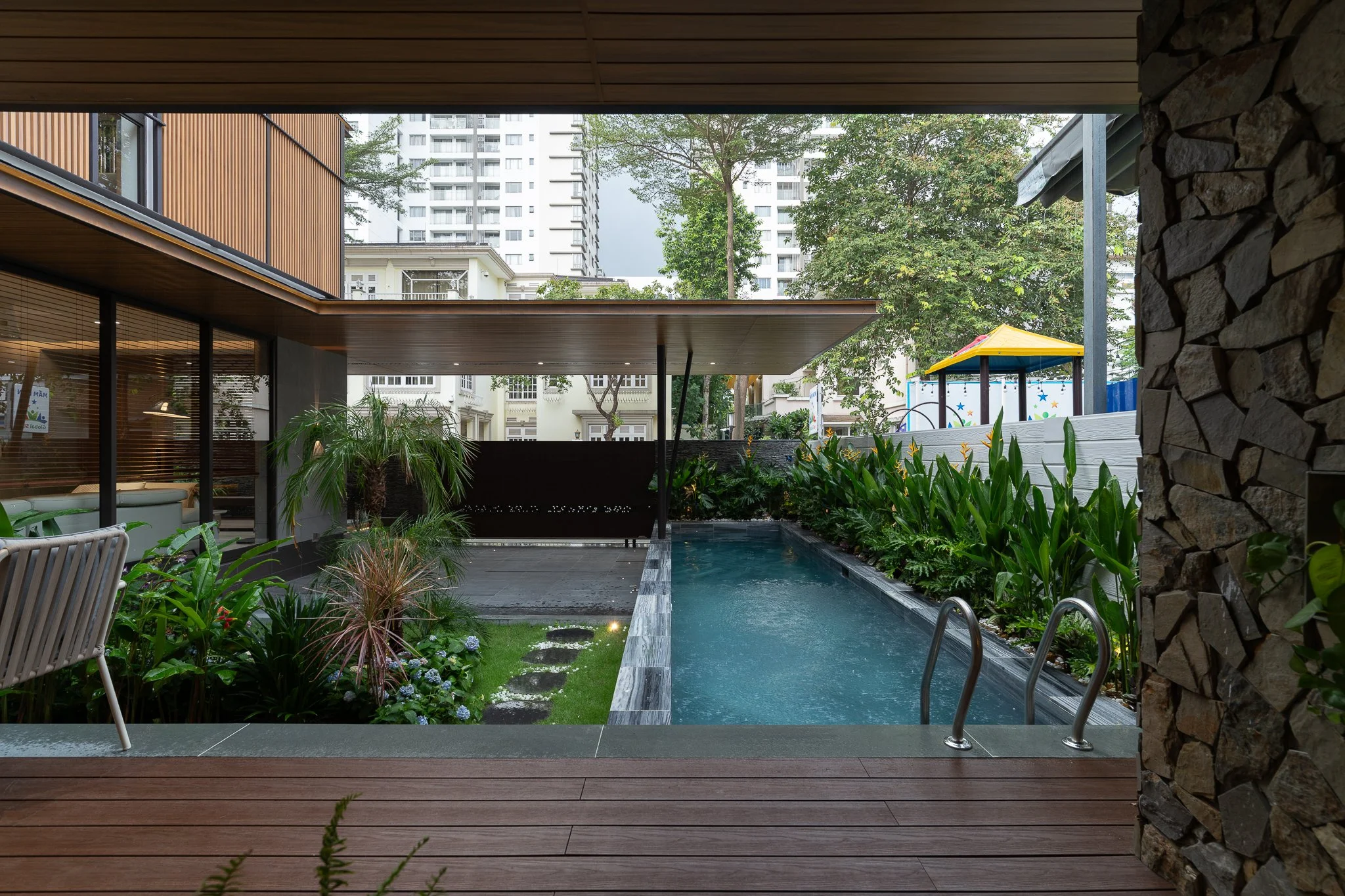 View of an outdoor swimming pool with surrounding plants, a house with large windows, and high-rise buildings in the background, seen from beneath a covered patio area.
