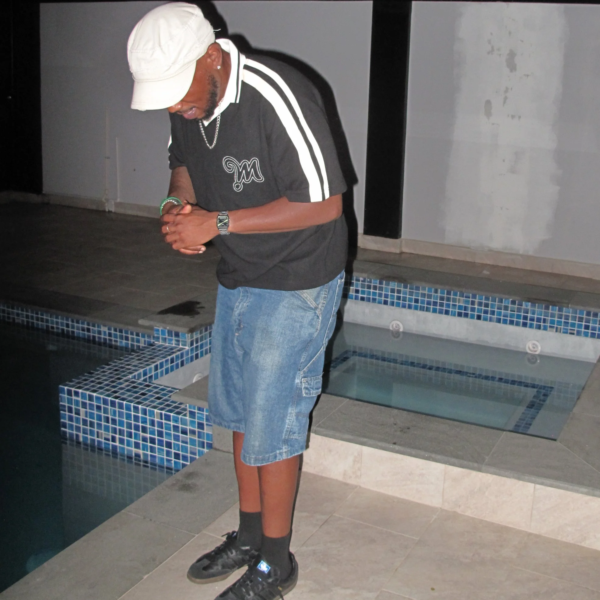 A man dressed in black with white stripes on his sleeves, wearing a white cap, glasses, a chain necklace, a watch, and colorful bracelets, is standing indoors near a tiled structure with a gray wall in the background.