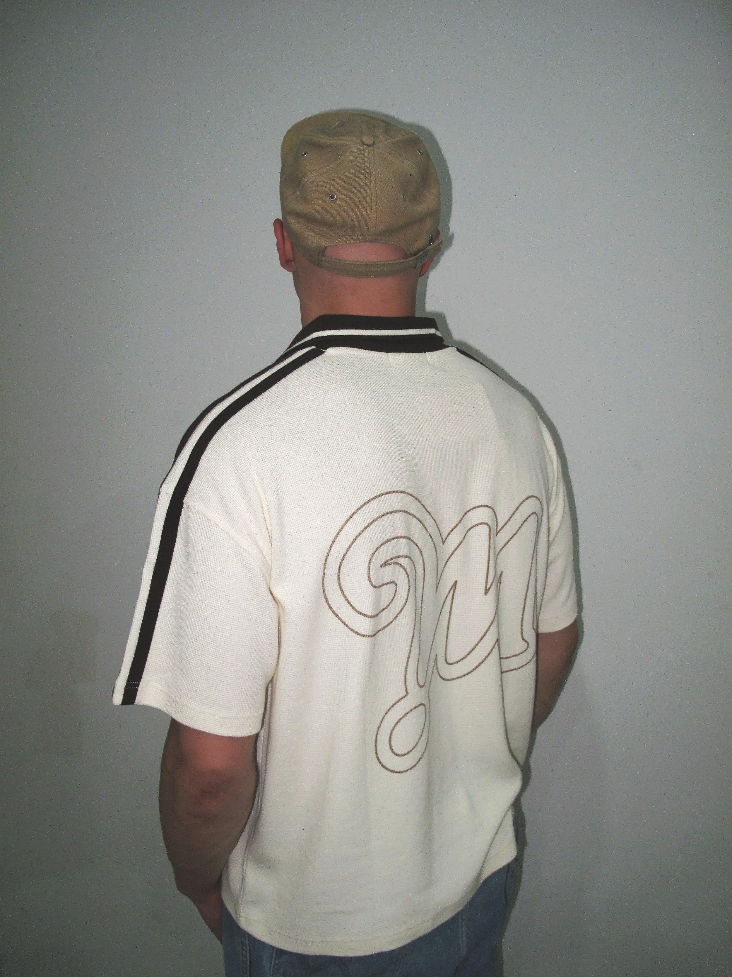 Back view of a person wearing a khaki cap and a white shirt with black stripes on the shoulders and large outlined text on the back, standing against a plain gray wall.
