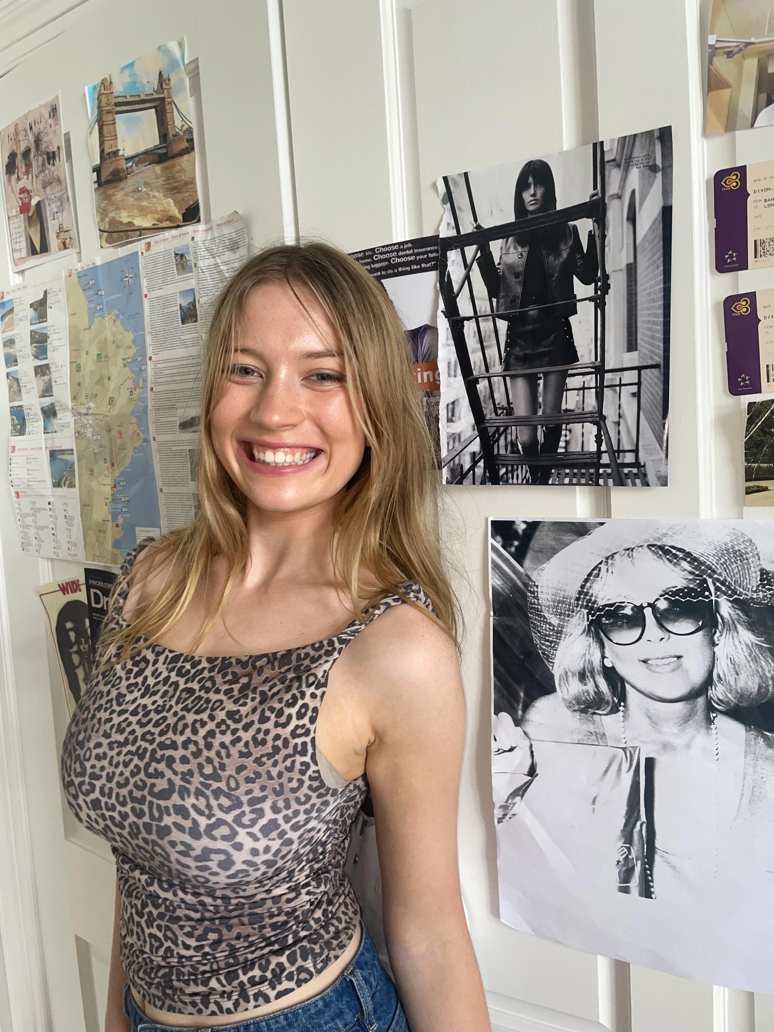 A young woman with blonde hair smiling, wearing a leopard print sleeveless top and blue jeans, standing in front of a wall with various posters and black-and-white photographs.