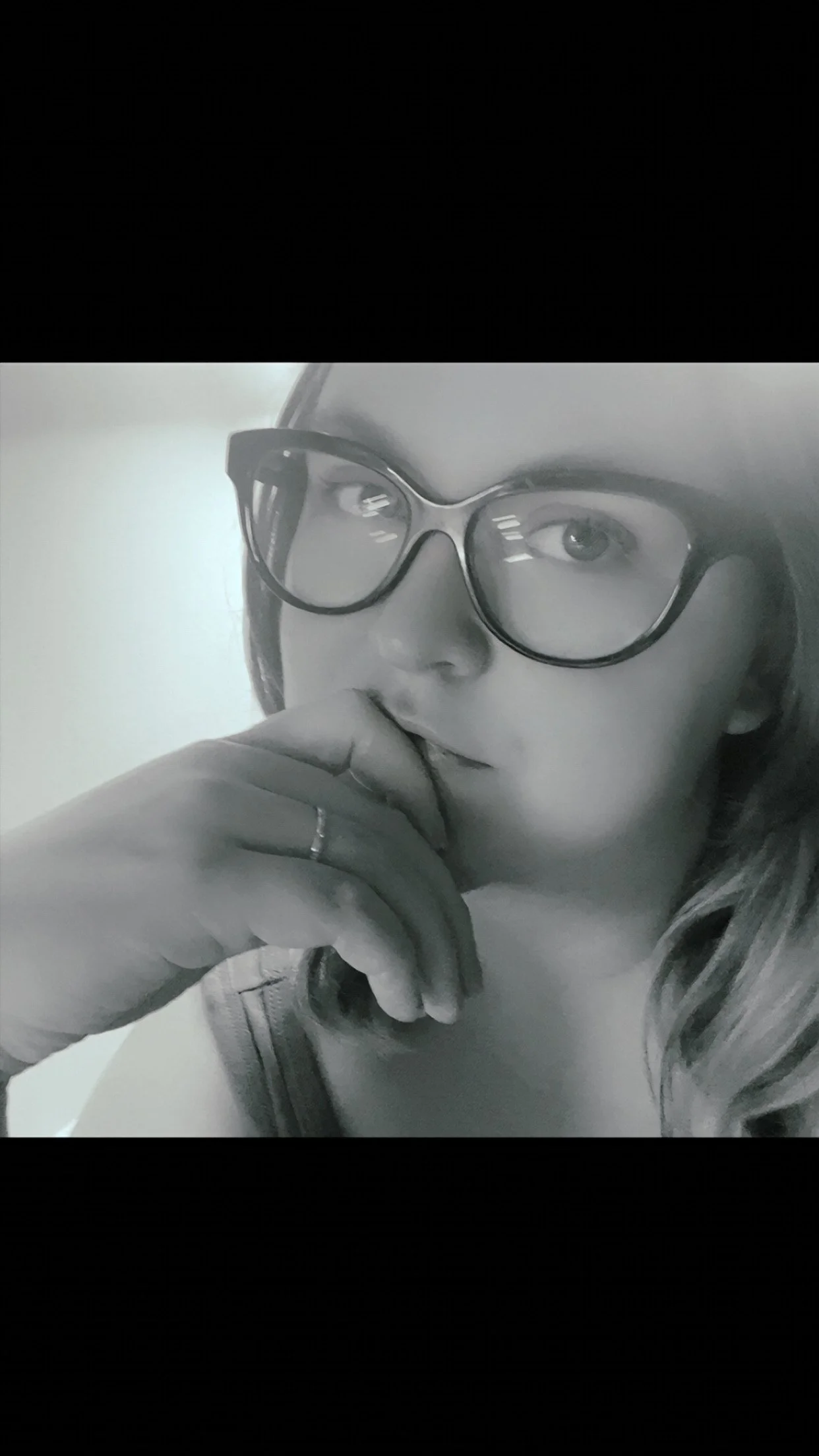 A black and white photo of a woman wearing large glasses, with her hand near her mouth, looking thoughtfully.