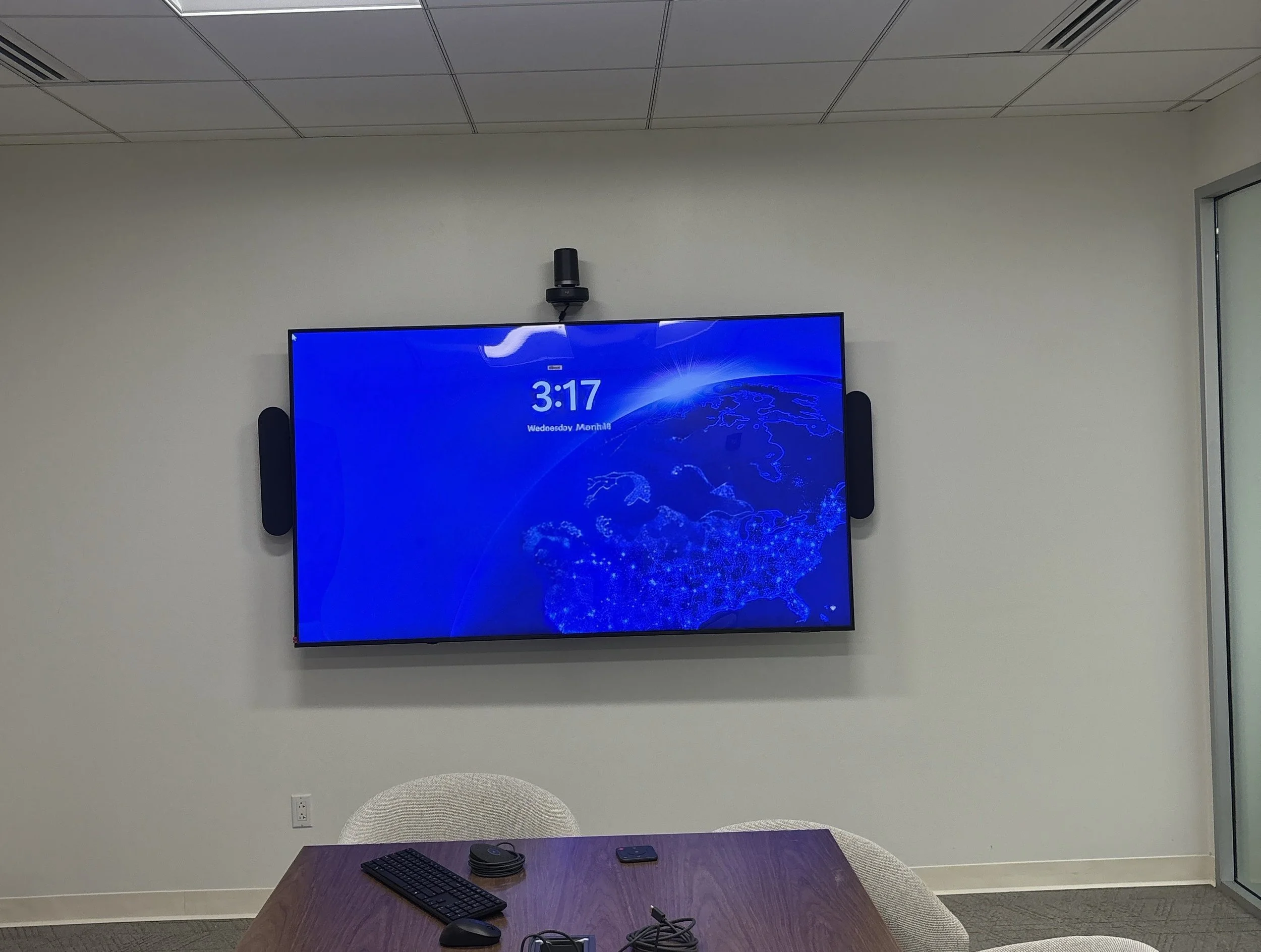 A conference room with a flat screen digital display showing the time as 3:17 and a world map background, mounted on a plain white wall. There is a small speaker above the screen, a table in the foreground with a keyboard, a mouse, a cellphone, and some cables, and a white chair with a rounded back in front of the table.