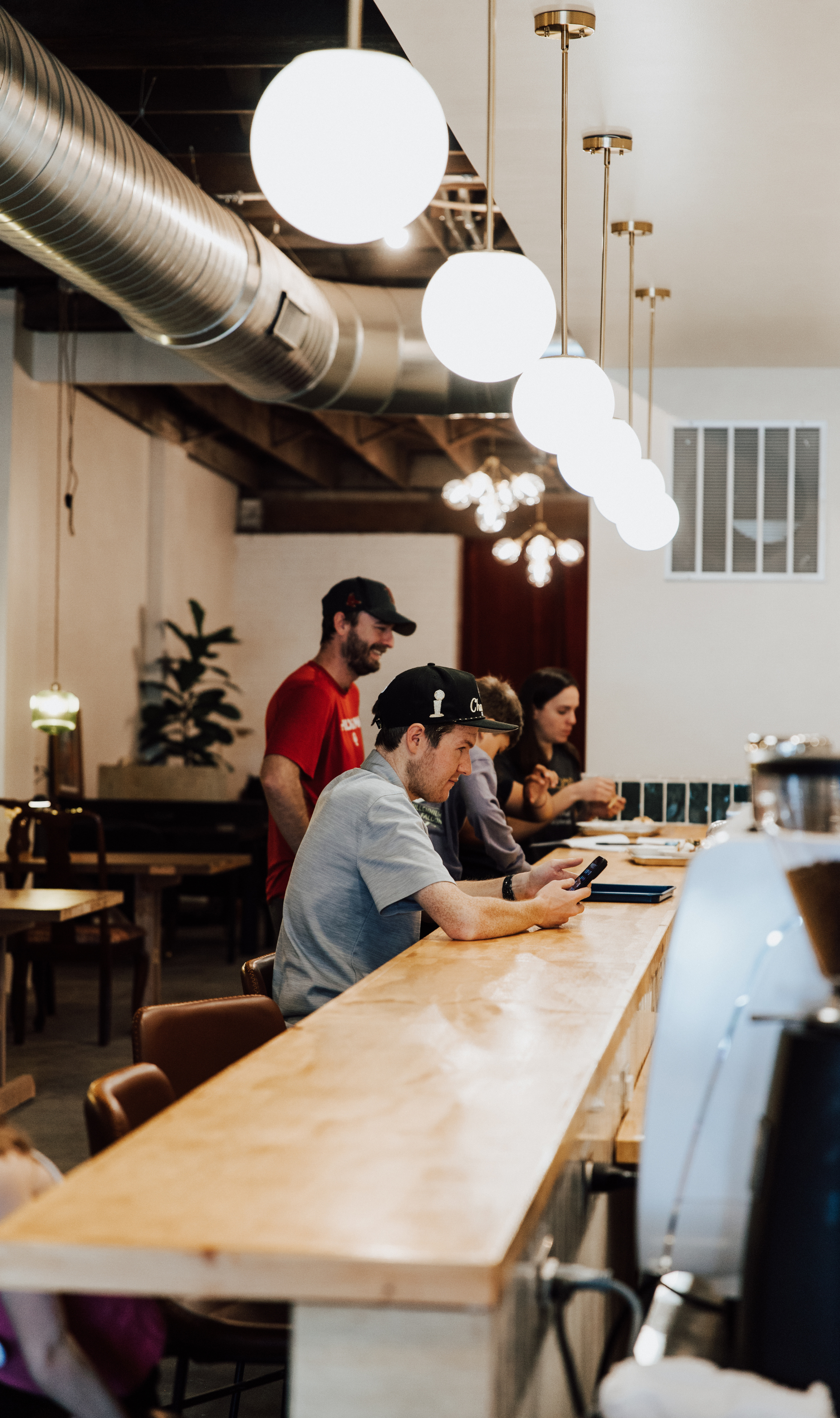 People sitting at a long wooden bar counter in a cafe, with contemporary hanging lights and industrial decor, including exposed ductwork and a ceiling with wooden beams.