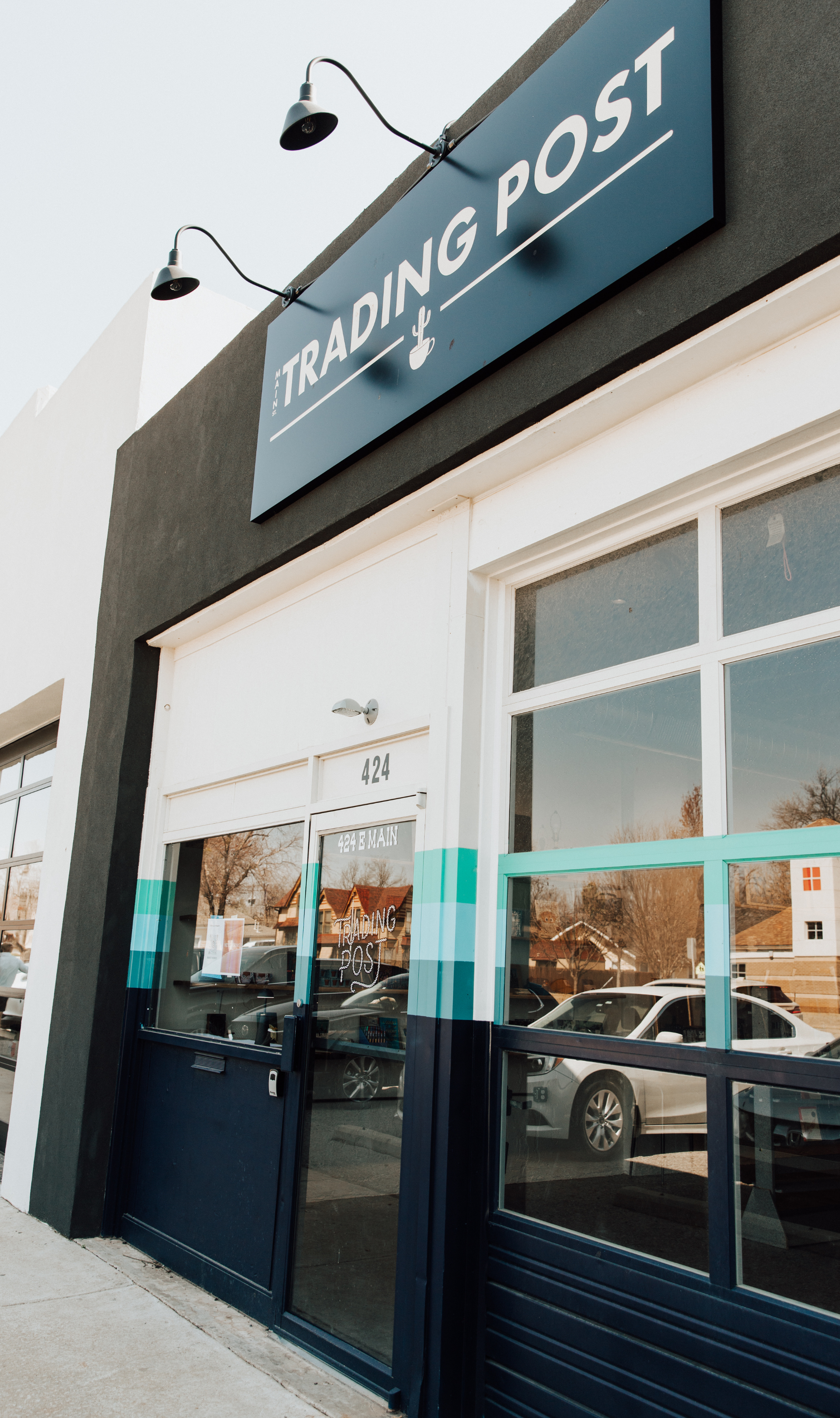 Exterior view of a trading post storefront with large glass windows, a dark-colored sign above with the text 'Trading Post' and a cactus icon, and two black outdoor lights mounted above the sign.