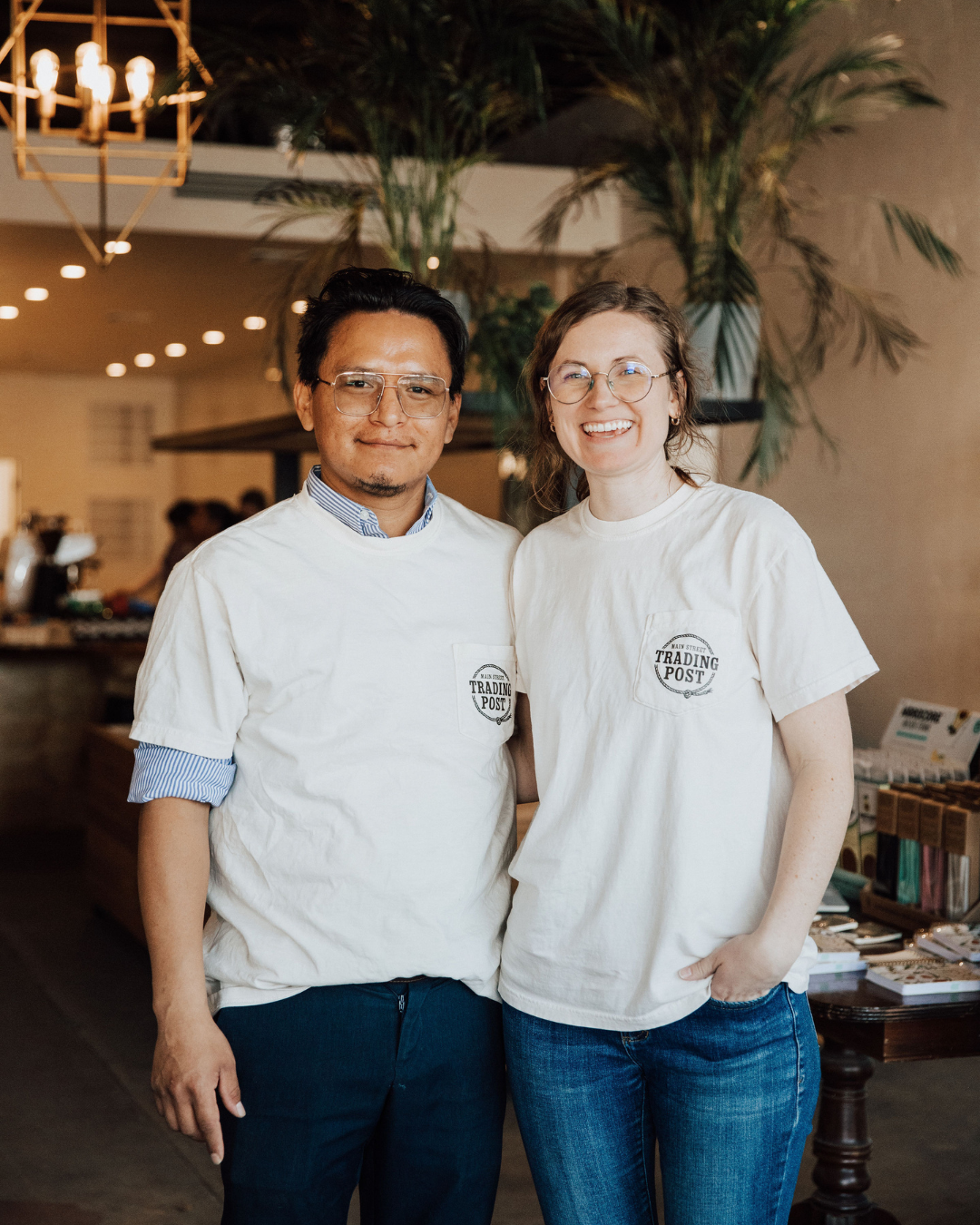 Two smiling people wearing white T-shirts with a logo that reads 'Trading Post' stand together indoors near a table with books and other items, with a large plant and warm lighting in the background.