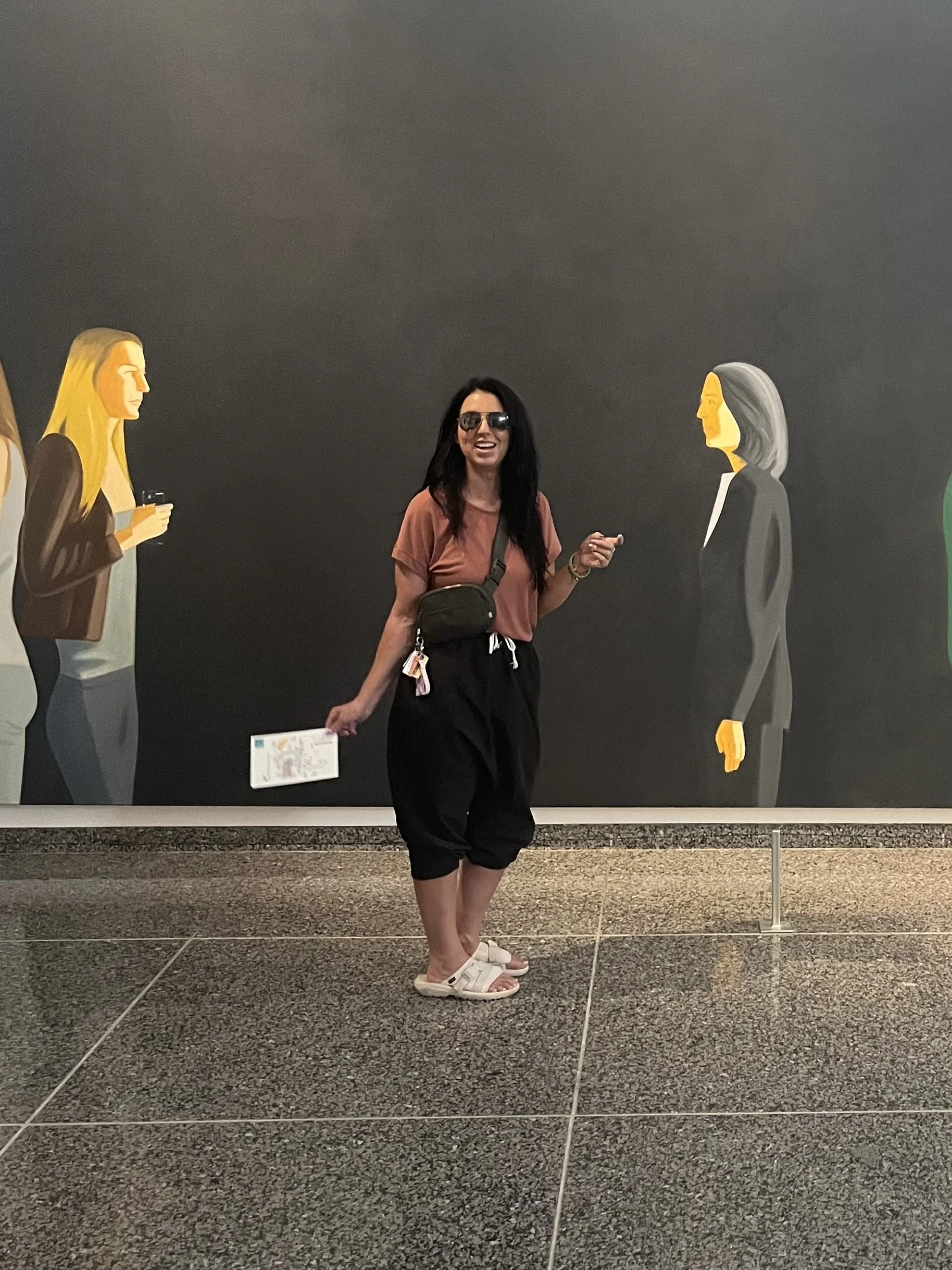 A woman with long dark hair, sunglasses, and casual clothing, smiling and pointing at something, standing in front of a dark mural with illustrated women.