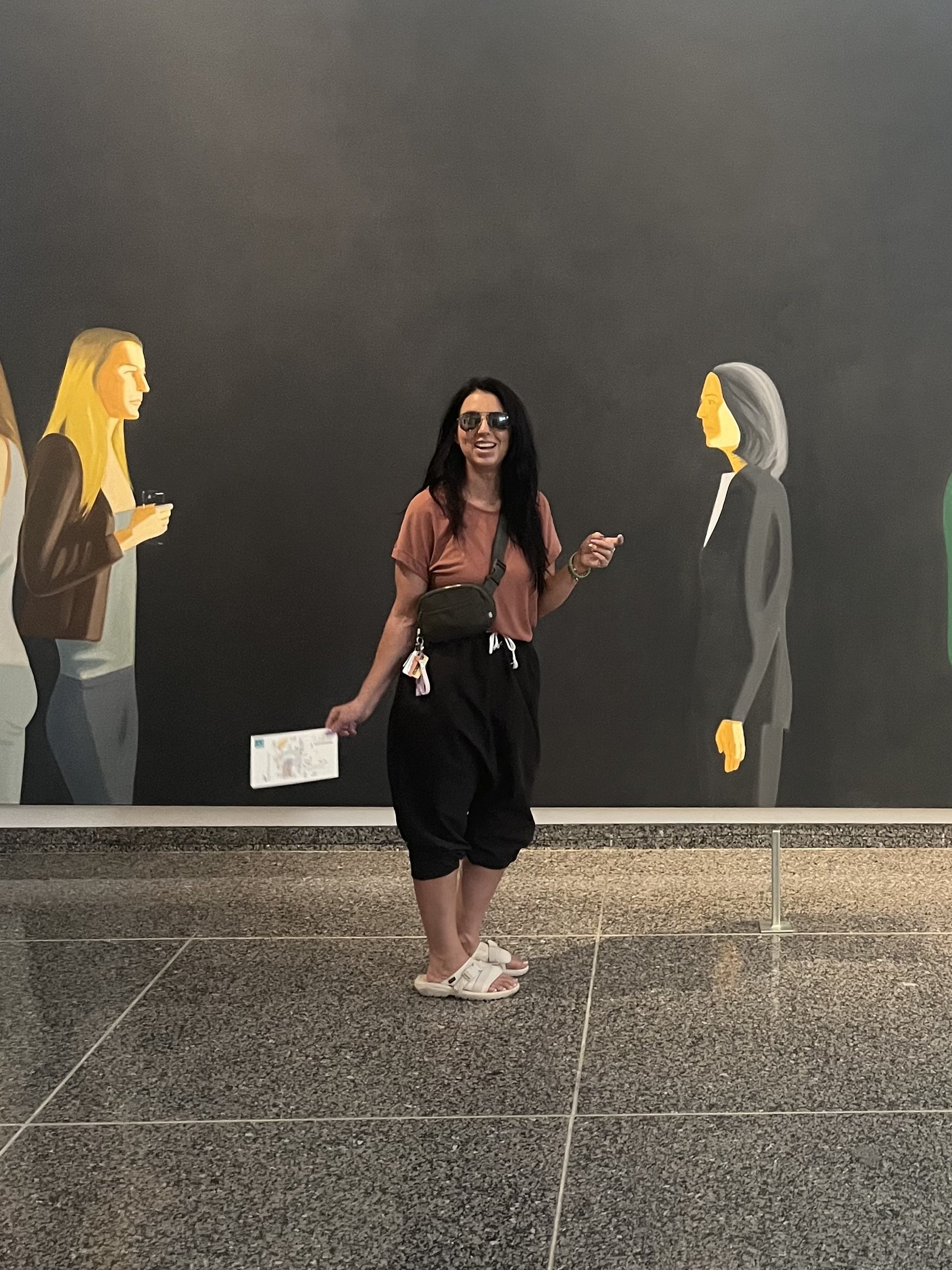 Woman standing in front of a mural of two stylized female portraits on a dark wall, wearing sunglasses, a peach T-shirt, black shorts, and white sandals, smiling and pointing.