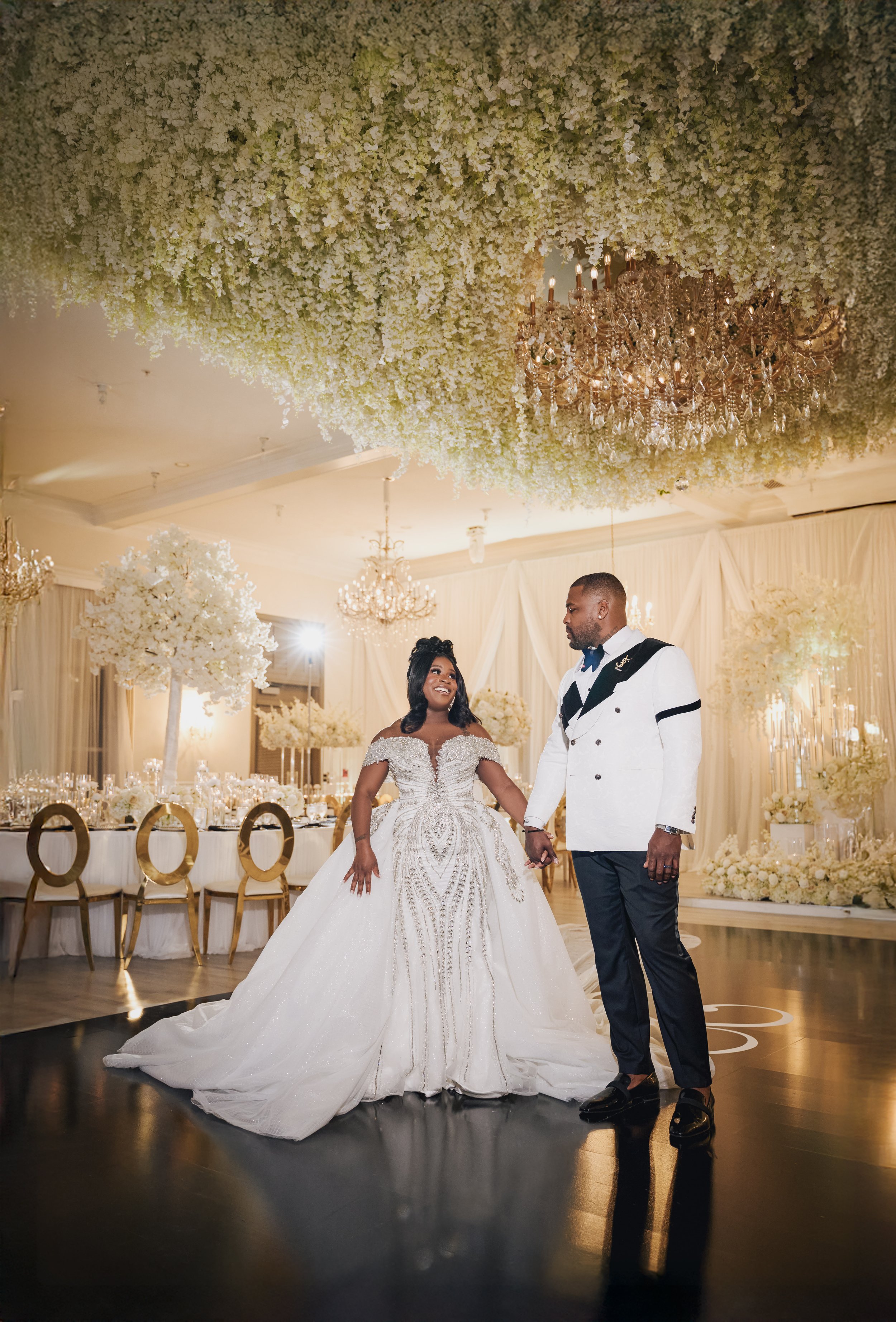 Bride and groom holding hands during their wedding ceremony, standing on a dance floor in an elegant banquet hall with chandeliers and floral decorations.