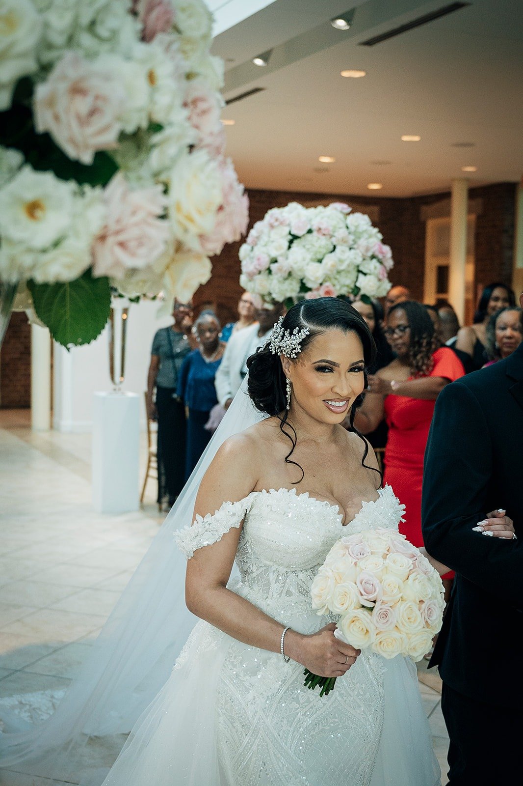 Bride in wedding dress holding a bouquet of light-colored roses, smiling, with guests and floral arrangements in the background.