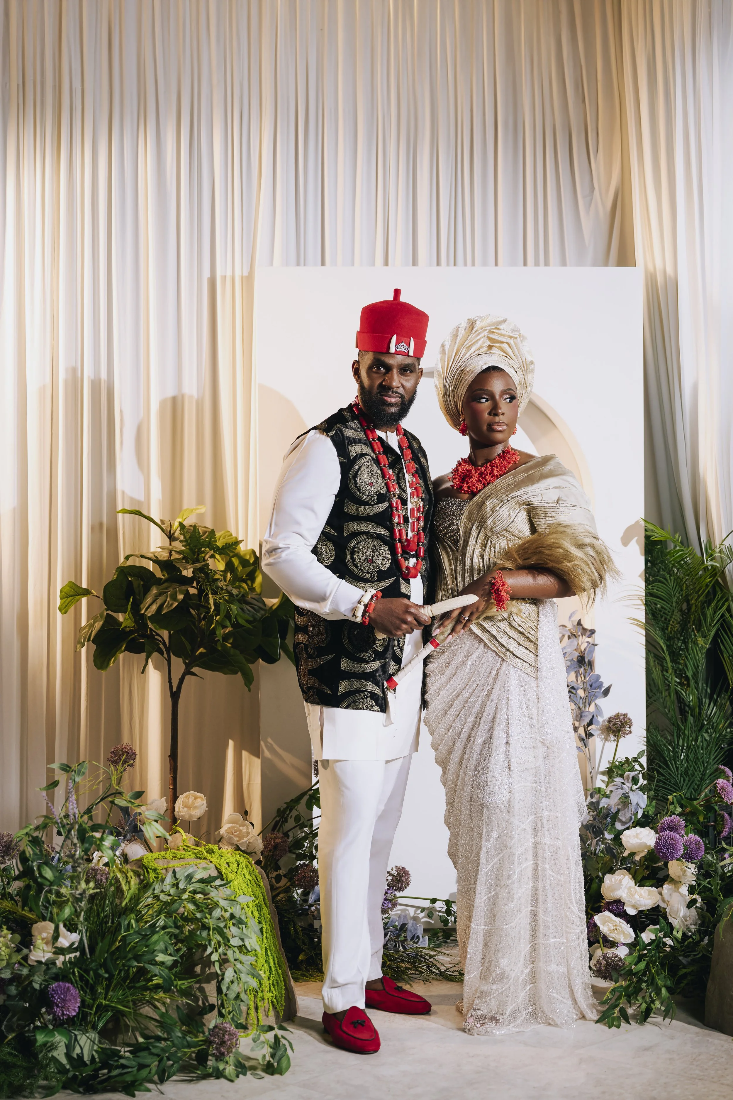 A man and woman dressed in traditional African attire standing at a wedding or special event, surrounded by flowers and greenery, with a light-colored curtain backdrop.