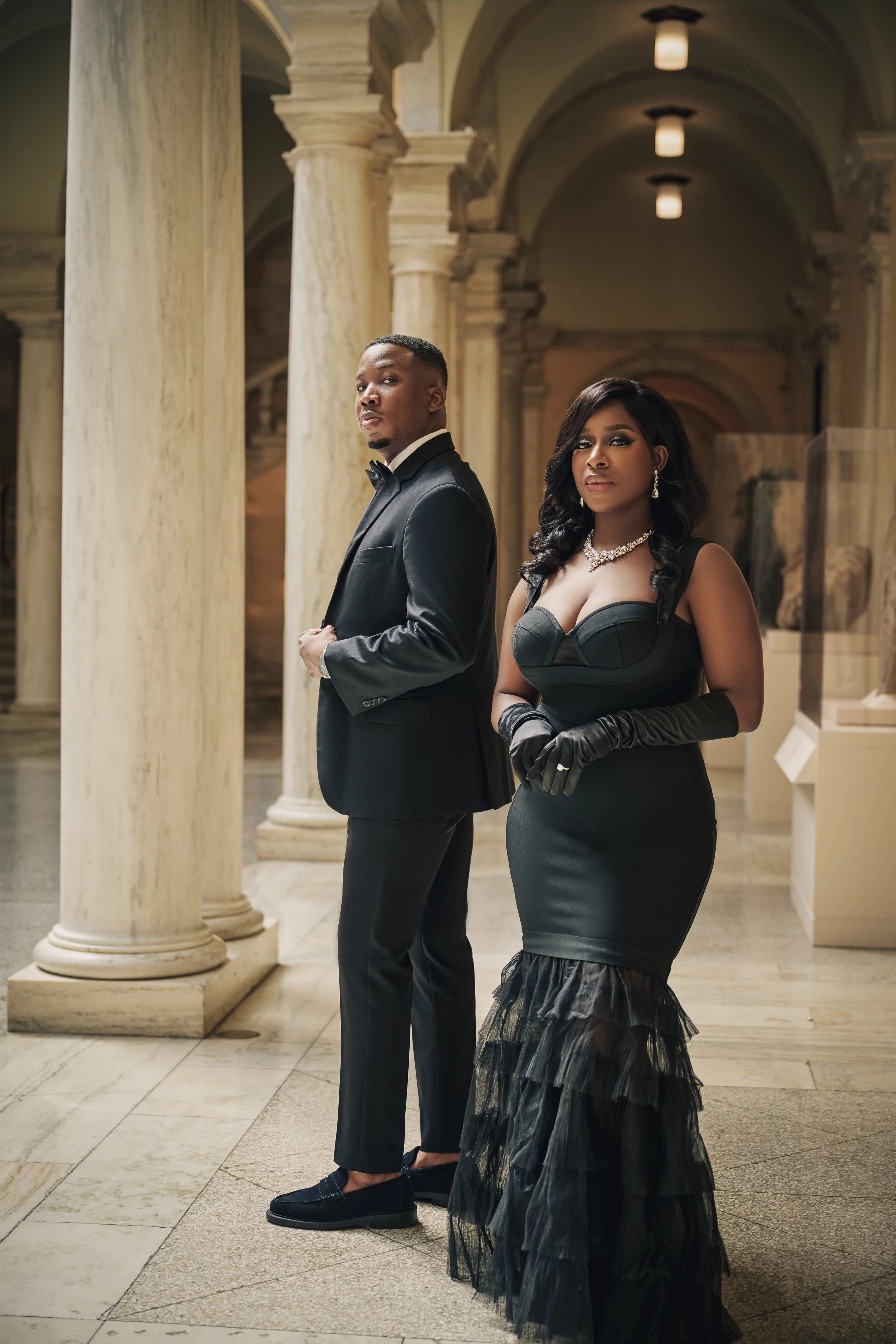 A well-dressed man and woman standing in an elegant, classical-style building with tall marble columns and arched ceilings. The man is in a black tuxedo with a bow tie, and the woman is in a black evening gown with gloves and jewelry.