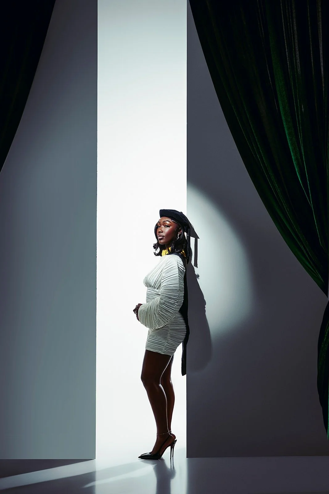A woman standing in a doorway with dramatic lighting, wearing a fitted dress with ruched details, high heels, and a black headpiece.