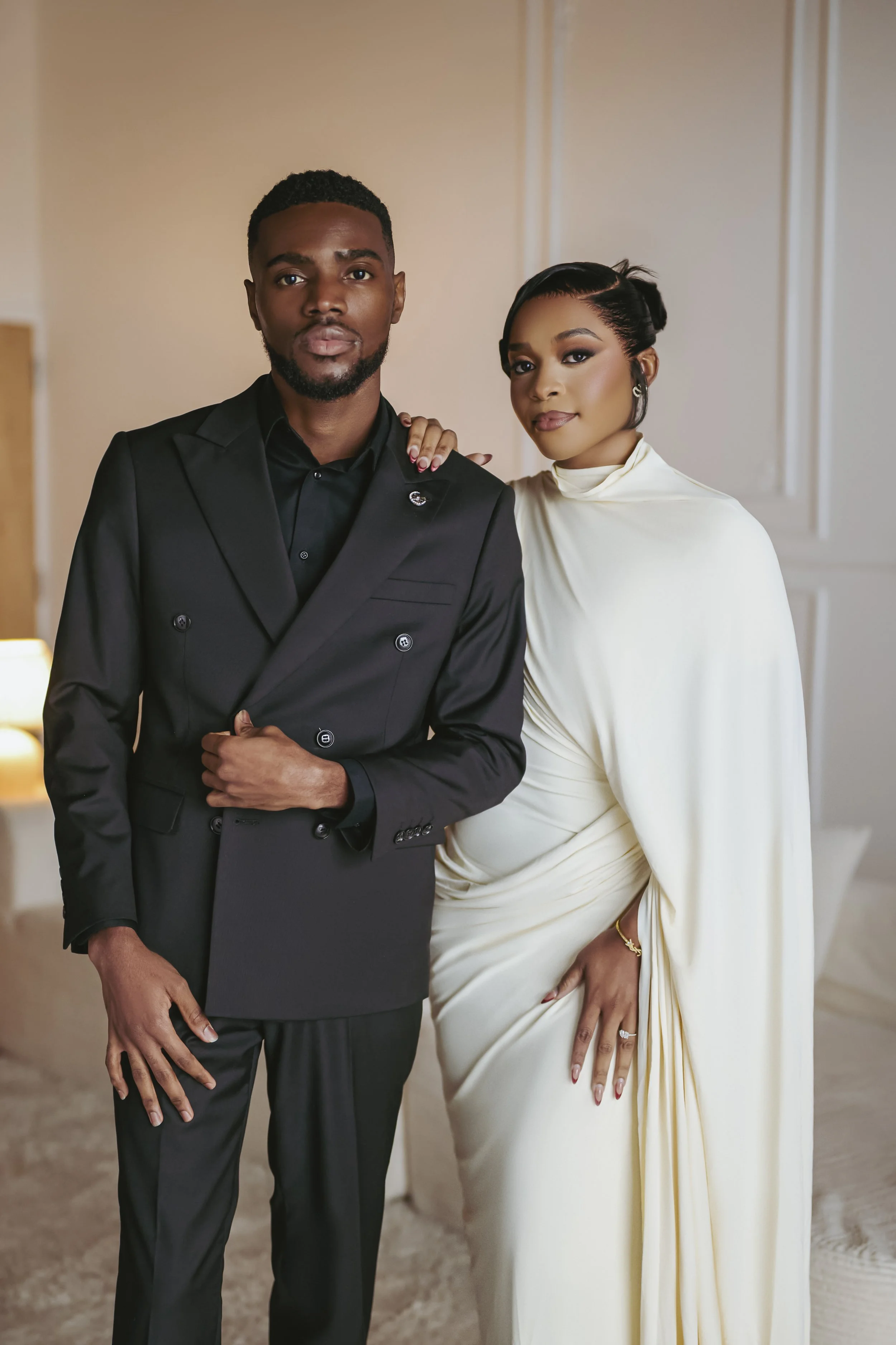 A well-dressed black man in a black suit and a white woman in an elegant cream-colored dress posing together indoors.
