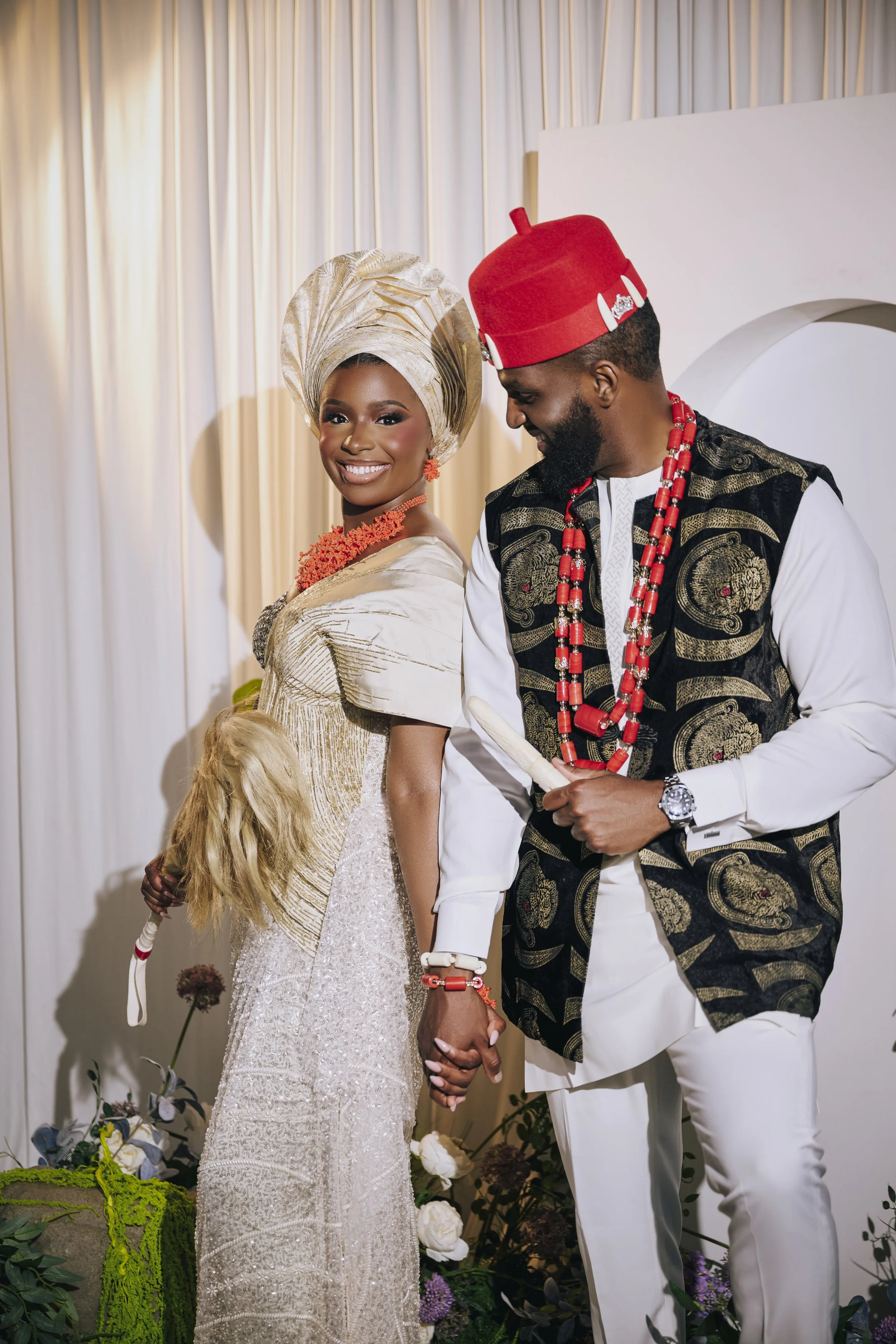 A couple dressed in traditional African wedding attire holding hands, standing in front of a floral decoration backdrop. The woman wears a gold crown and matching jewelry, the man wears a red hat and patterned vest with beaded necklaces.