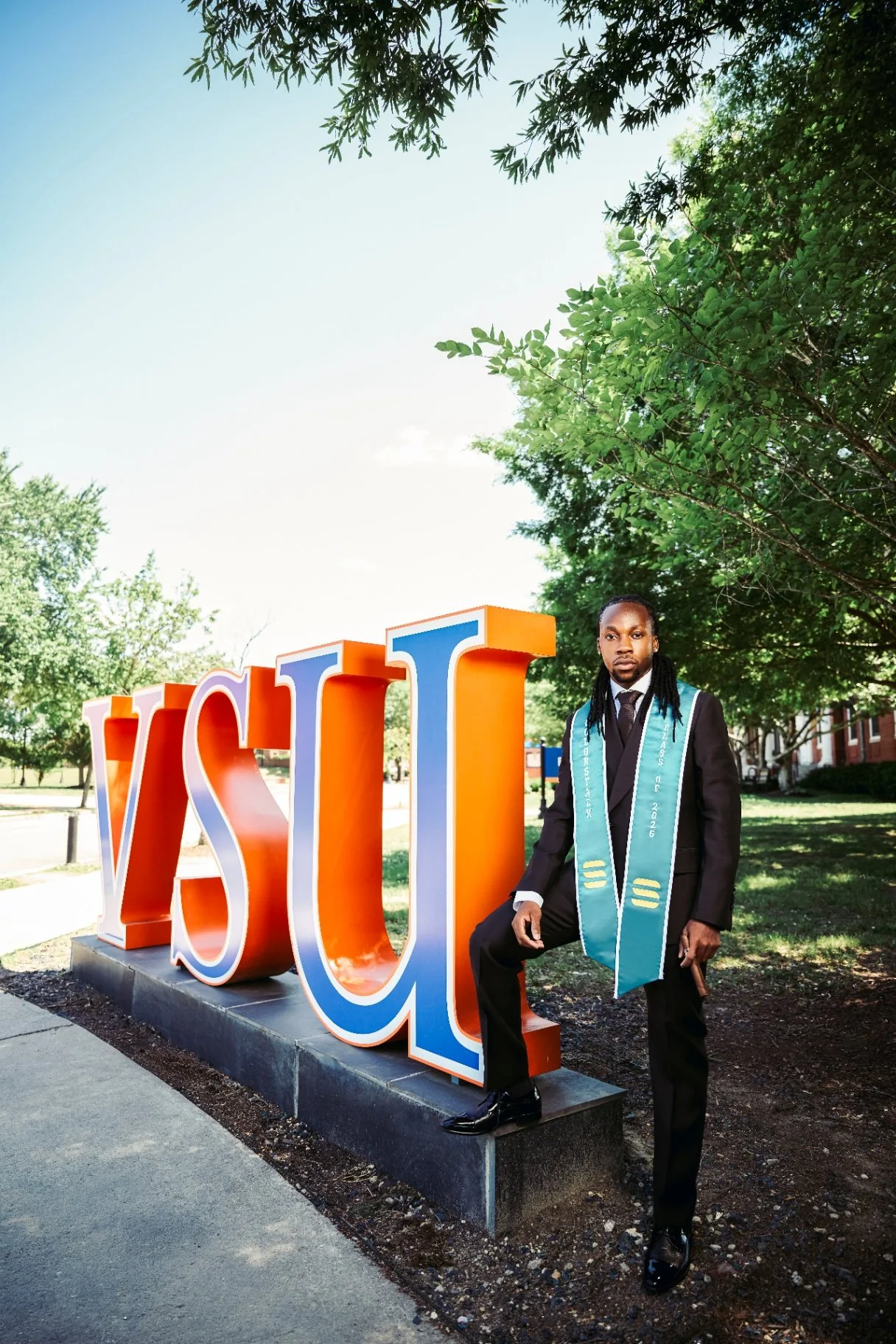 A man in a black suit with a graduation stole sits on a large university sign that reads "VSU" outdoors on a sunny day, with trees in the background.