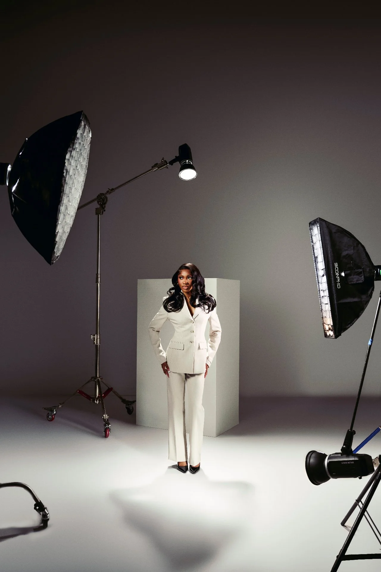 A woman in a white suit poses in a photography studio with professional lighting equipment around her.