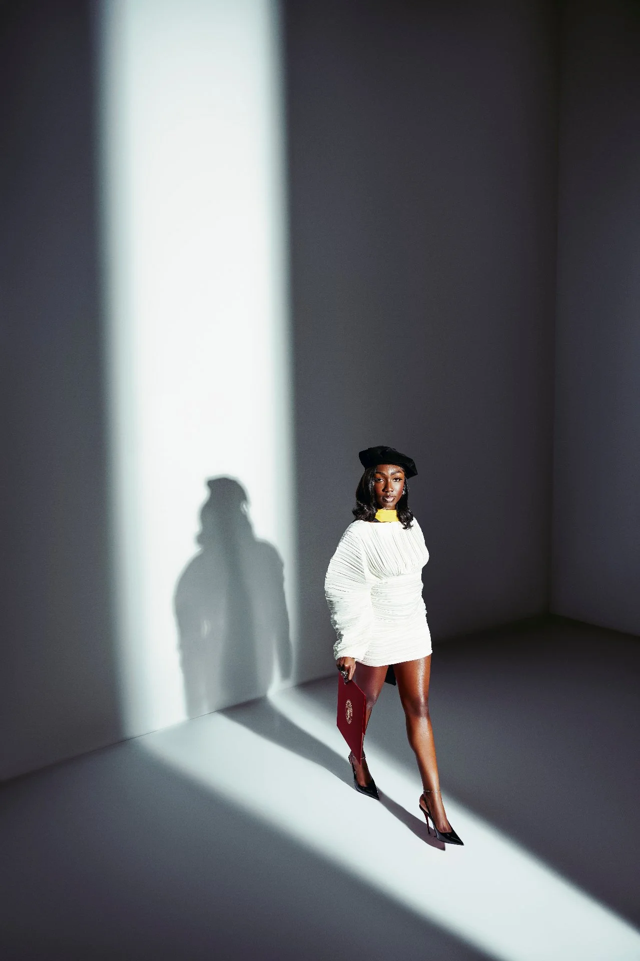 A woman standing in front of a plain wall, with dramatic lighting casting her shadow and a secondary shadow of her profile on the wall. She is wearing a white dress with a yellow collar, a black hat, and high heels, holding a red folder in her hand.