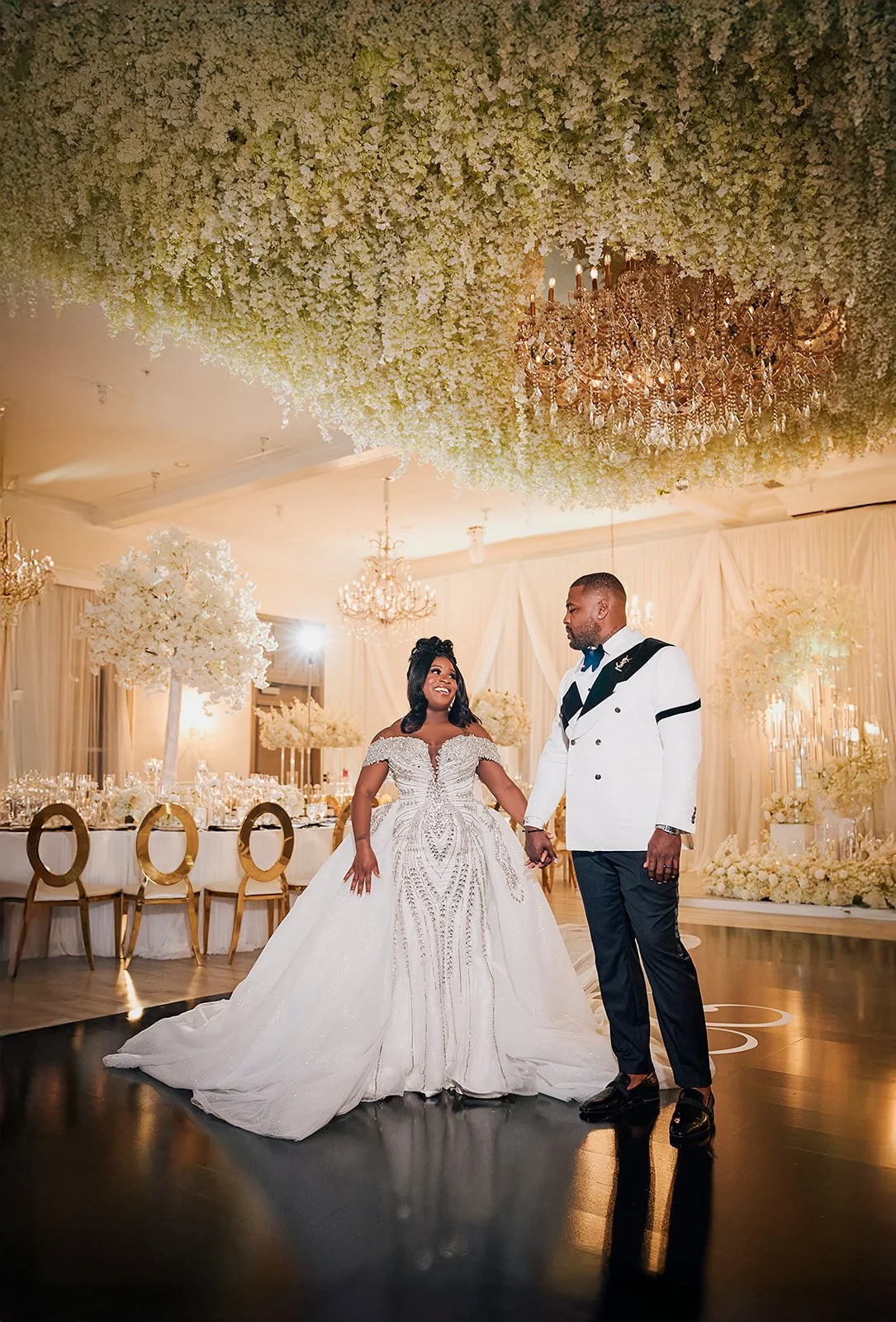 A bride and groom holding hands at their wedding reception, surrounded by elegant floral arrangements and chandeliers in a decorated banquet hall.
