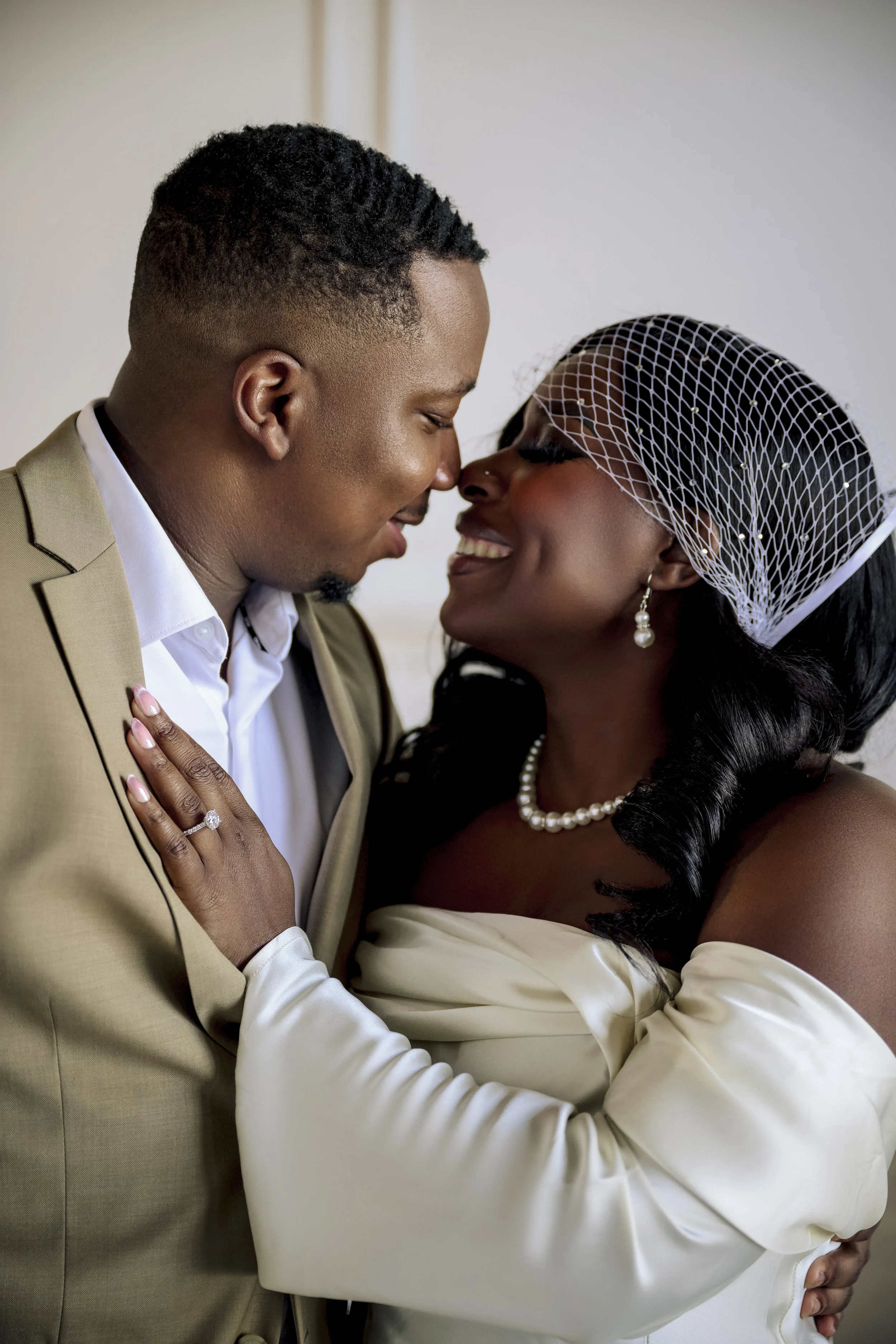 A couple on their wedding day, nose to nose, smiling, with the woman wearing a veil and pearl jewelry, and the man in a beige suit, in an intimate pose.