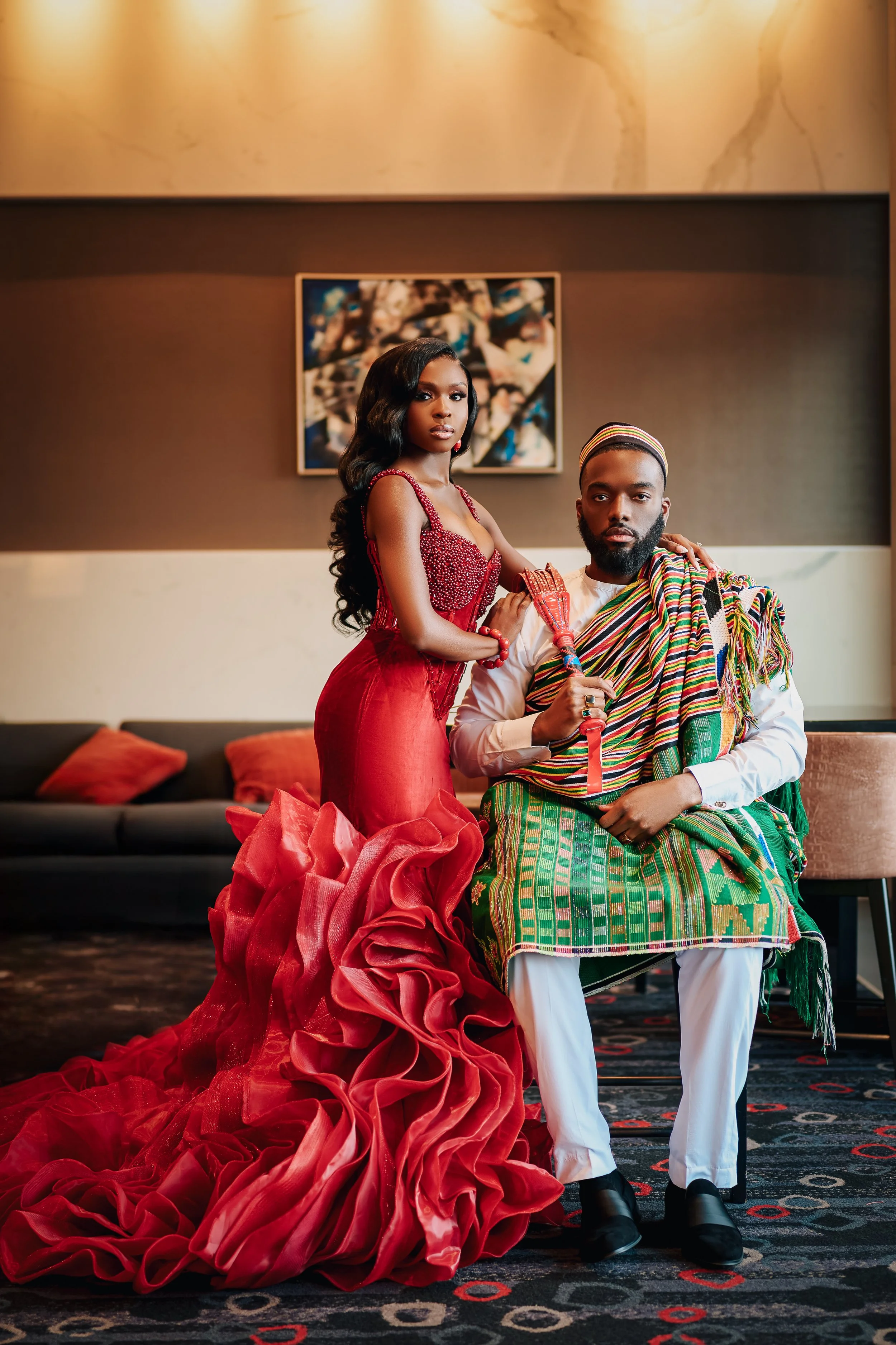 A woman in a red dress with ruffles and a man dressed in traditional African attire pose indoors. The woman has long black hair, and the man has a beard and a headband. They are sitting on a sofa with red cushions, with a dark-colored wall and an abstract painting in the background.