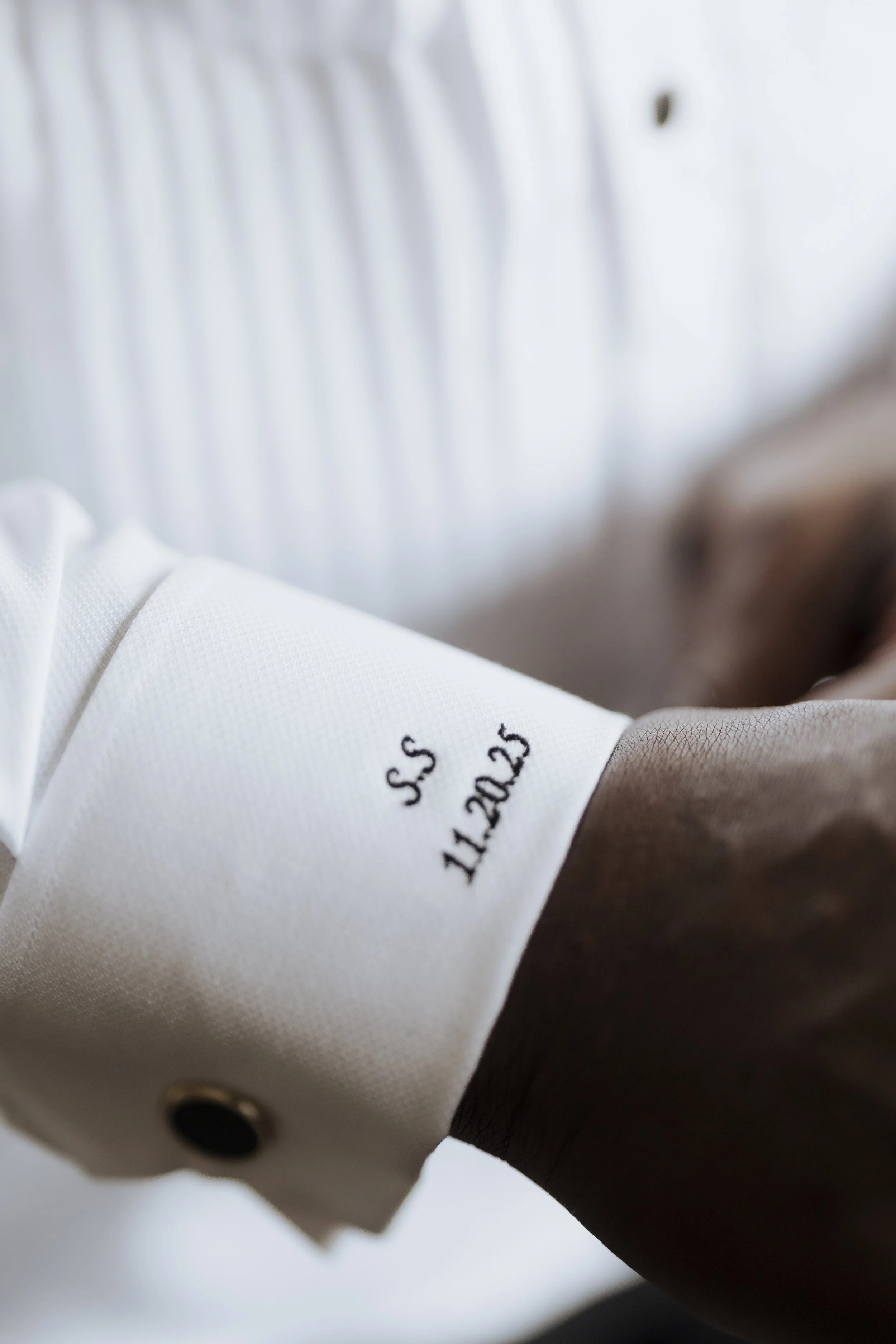 Close-up of a person in a white shirt with black buttons, showing a wristband with printed text, against a blurred white background.