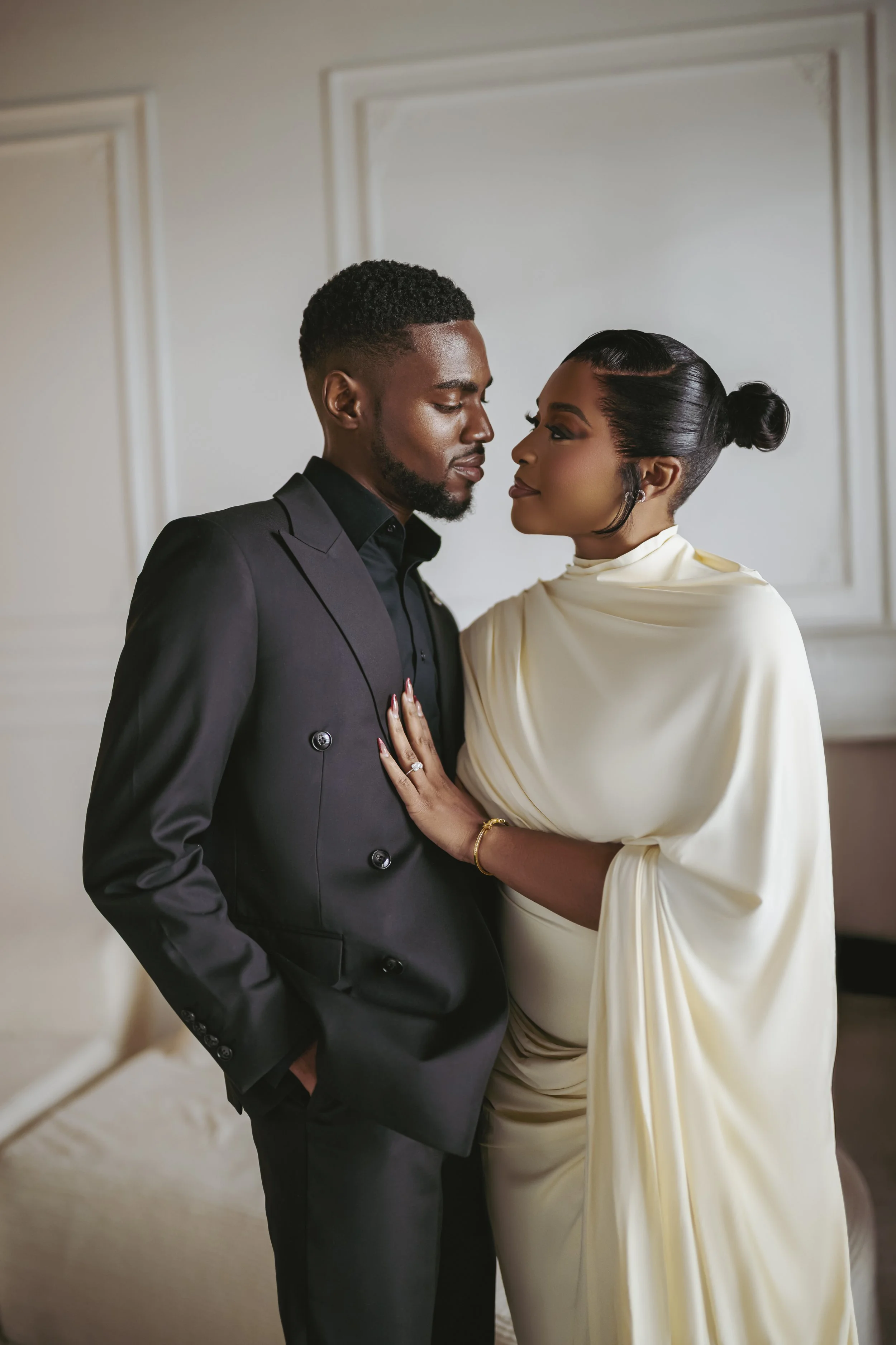 A man and woman in formal attire face each other closely, with the woman touching the man's chest and both gazing at each other affectionately, in an elegant interior setting.