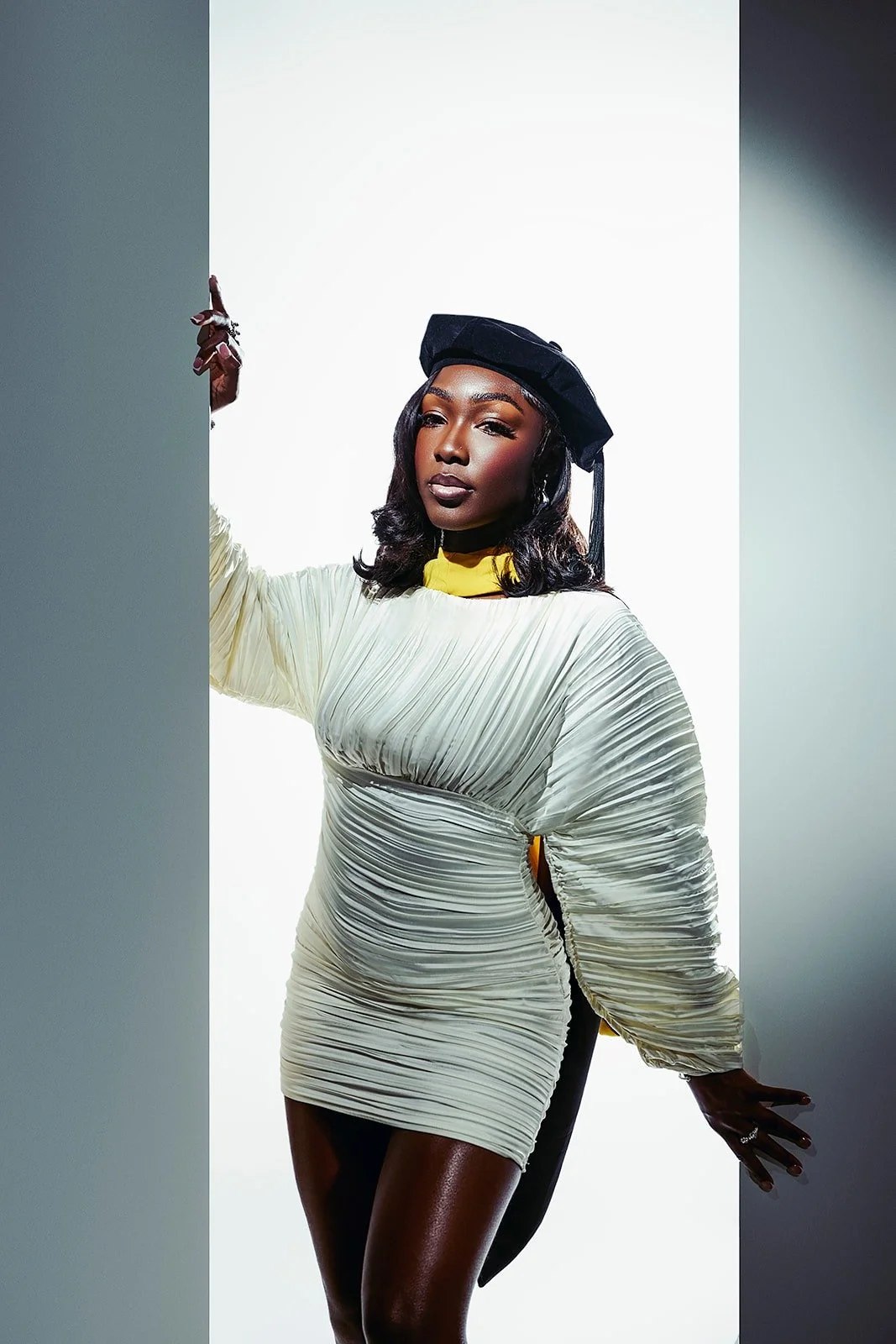 A confident woman with dark skin, wavy black hair, and wearing a black graduation cap stands against a white background, dressed in a shiny, textured cream-colored dress with a yellow collar.