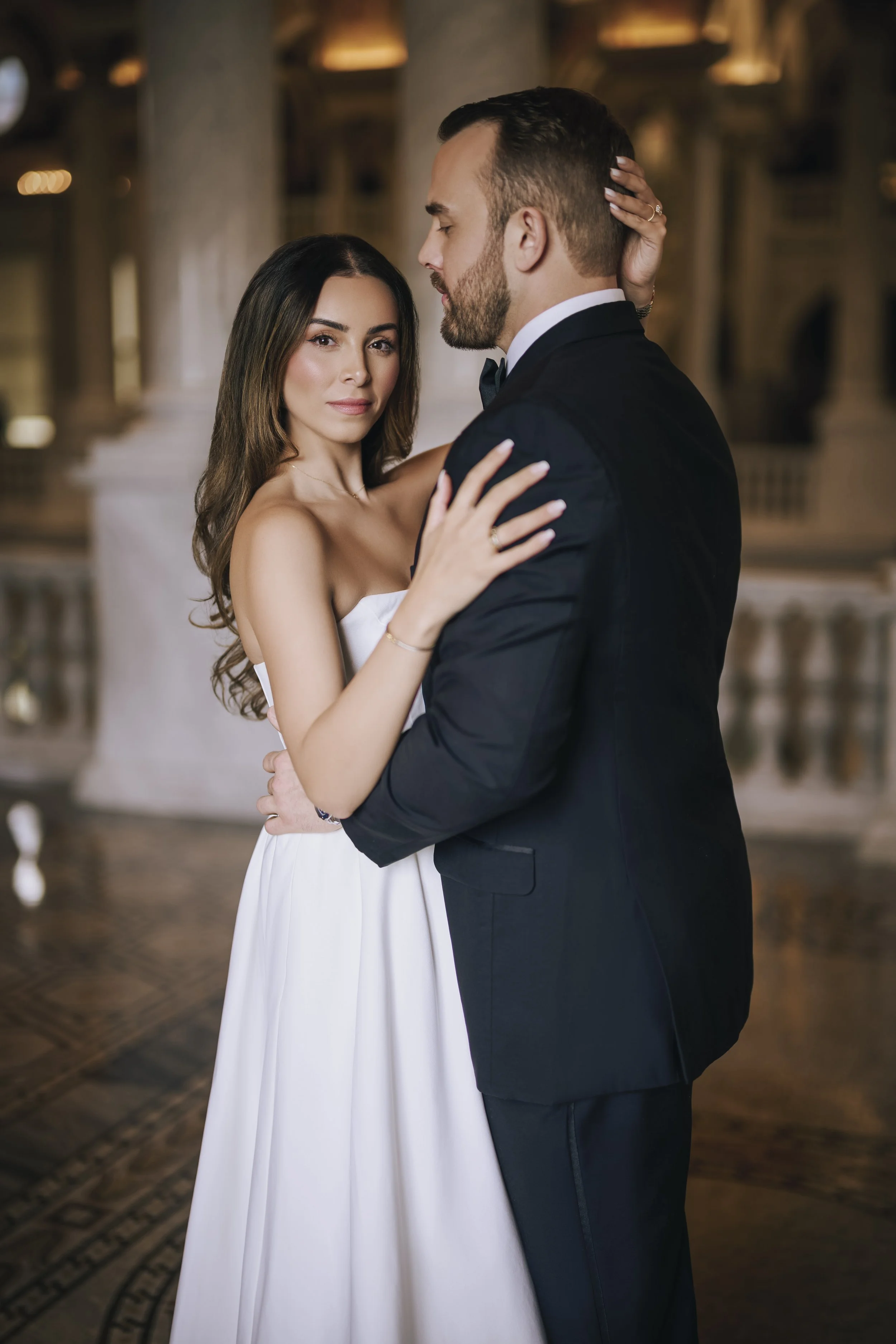 A couple dancing closely in formal attire, with the woman wearing a strapless white dress and the man in a black tuxedo, in an elegant, historic setting.