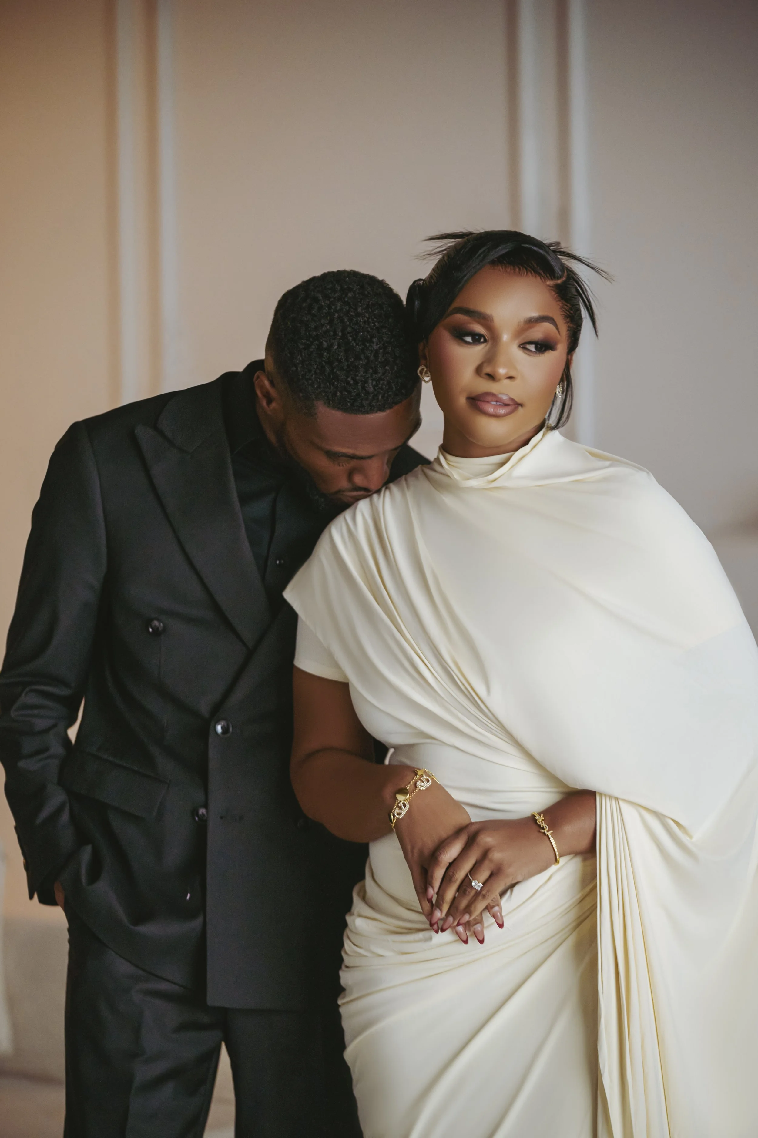 A couple dressed in formal attire, the man in a black suit with a boutonniere and the woman in an elegant cream-colored dress with jewelry, posing together.