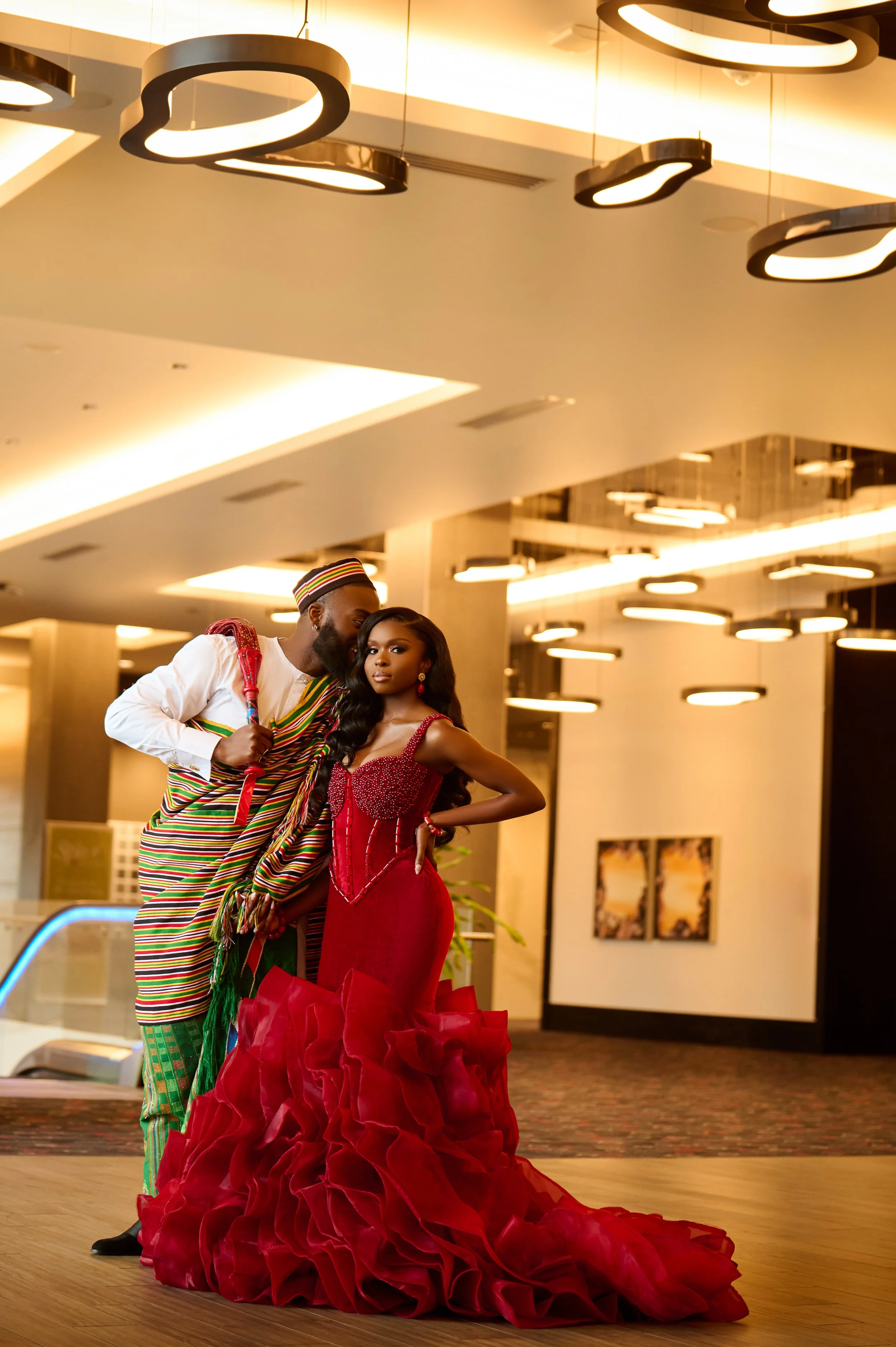 A man and woman in formal attire standing close together in an indoor setting with modern lighting and decor. The woman is wearing a red gown with ruffles, and the man is dressed in a colorful traditional African outfit.