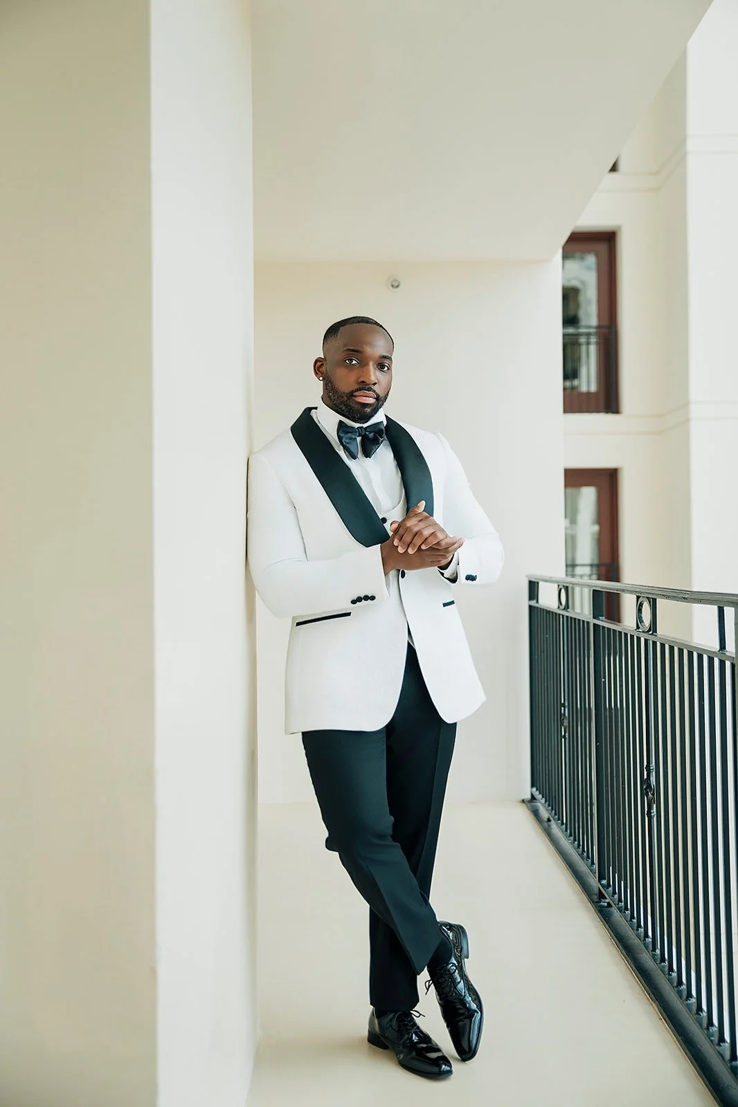 A man dressed in a formal white tuxedo jacket with black accents, black bow tie, black pants, and shiny black shoes, leaning against a white wall on a balcony, with modern building windows in the background.